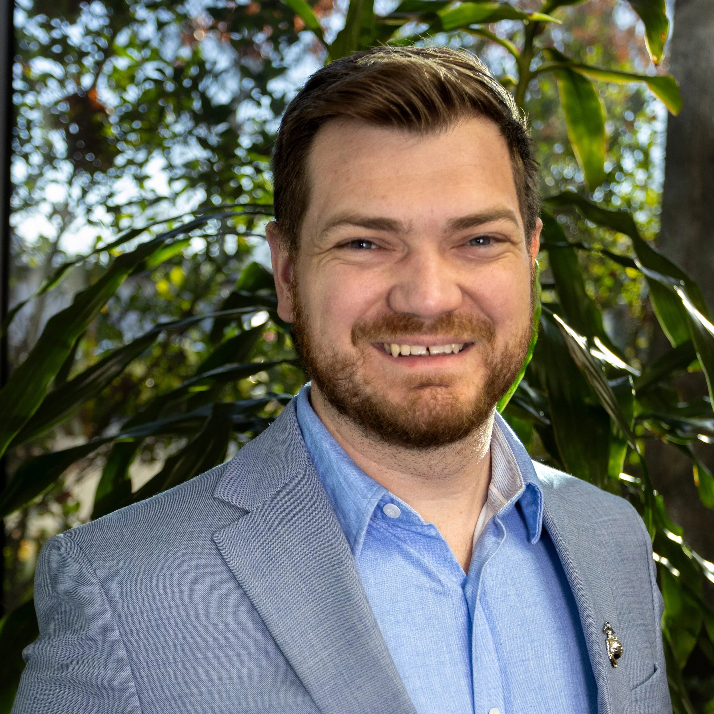 A man with short brown hair, a beard, and a mustache, wearing a light gray blazer and a light blue dress shirt, smiling outdoors with green foliage in the background.