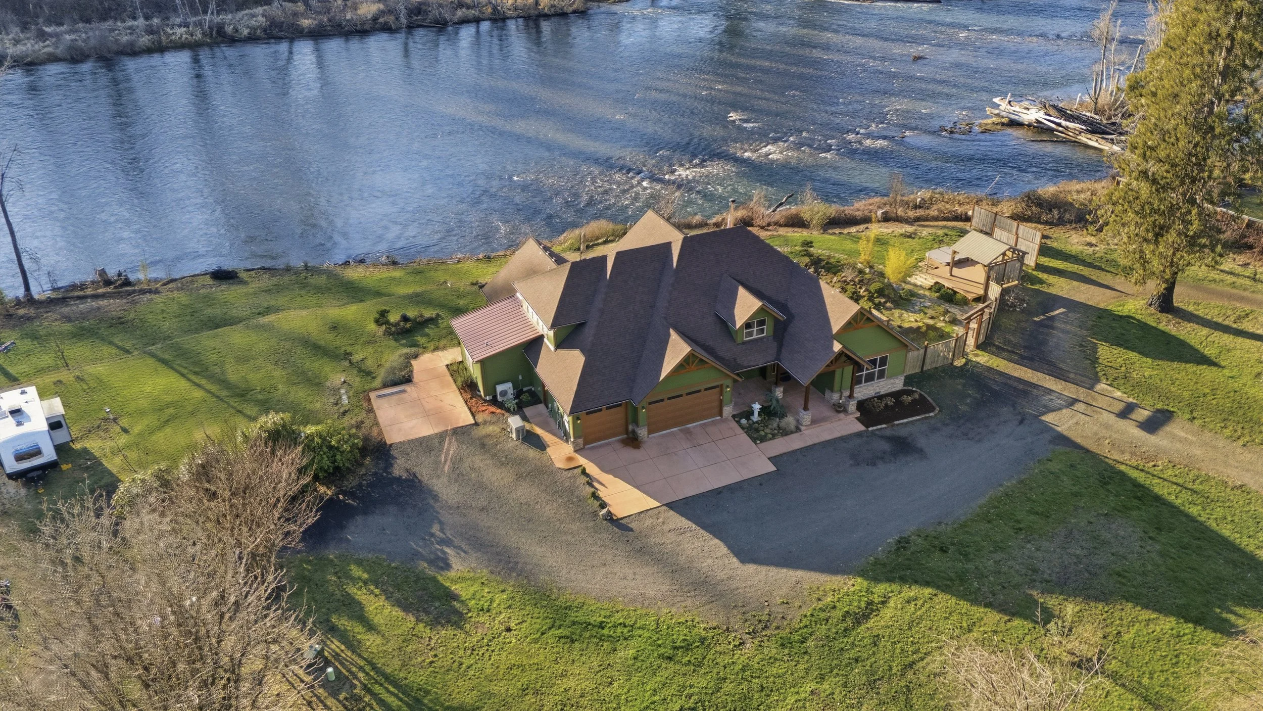 Aerial view of a house near a body of water with a lawn, driveway, and surrounding trees.