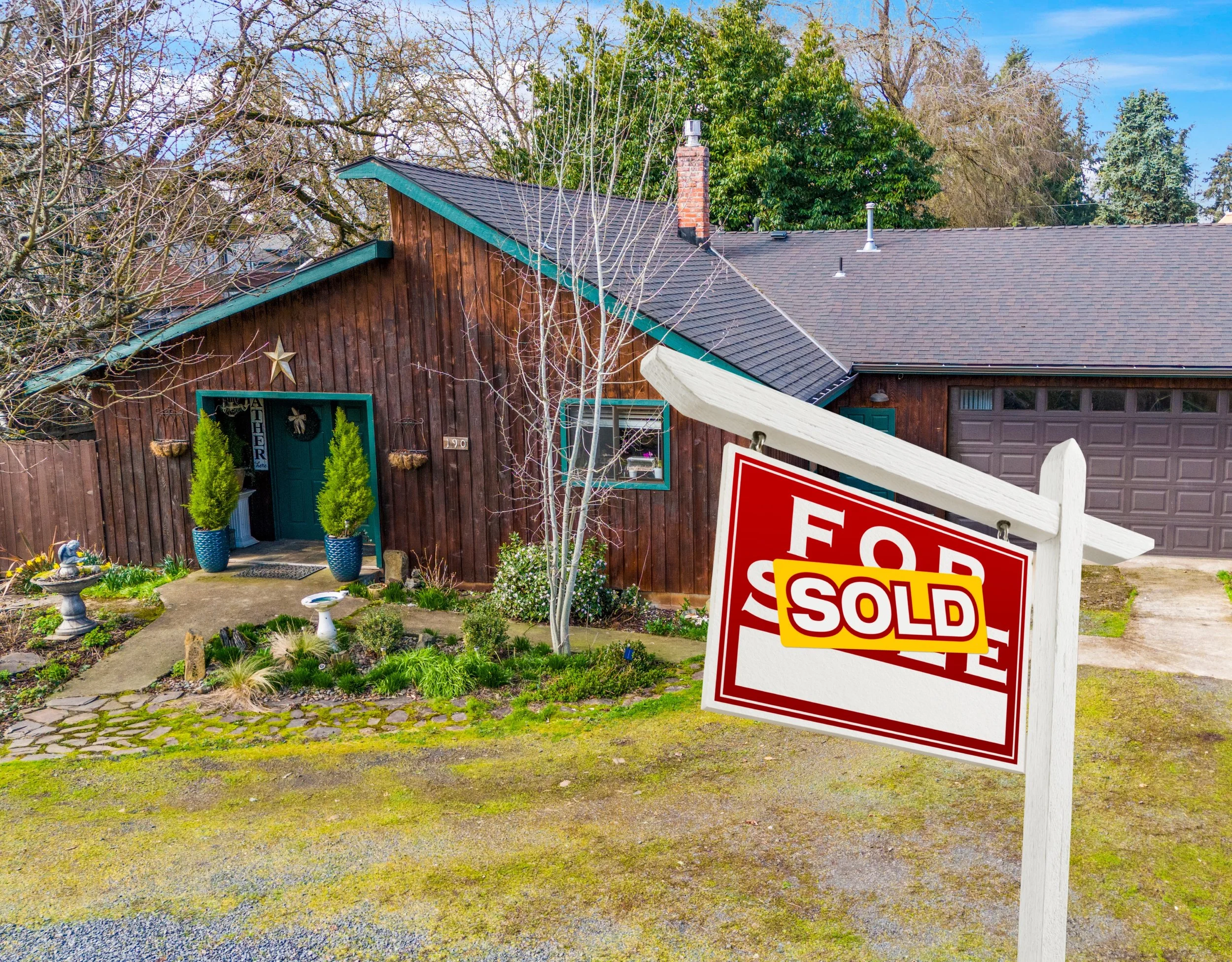 A house with dark wooden siding, green trim, and a sloped roof, with a 'Sold' sign in the front yard indicating the property has been purchased.