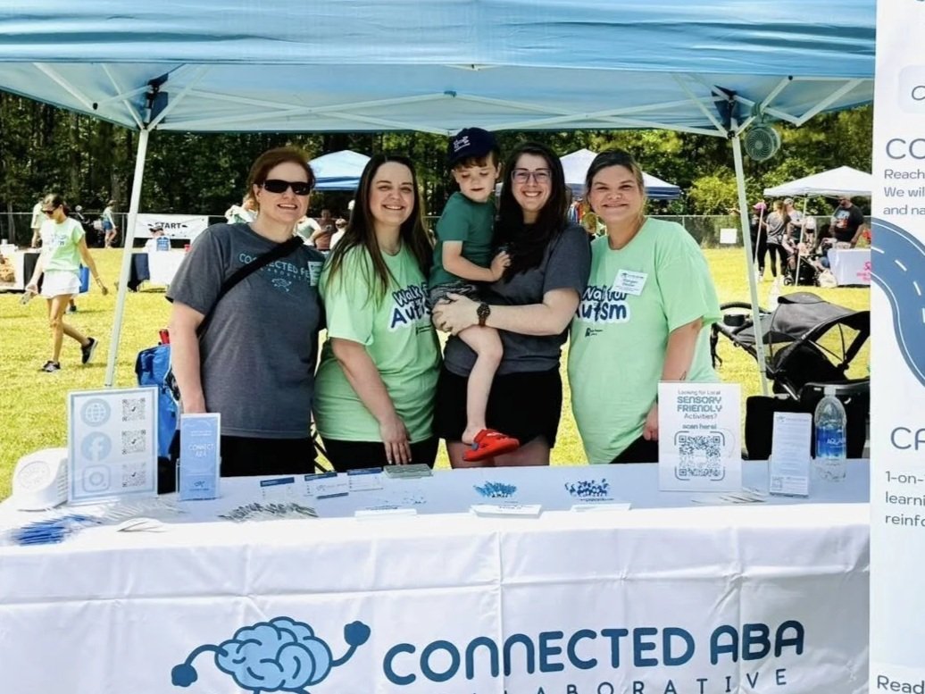 Group of five people standing behind a table at an outdoor event, under a blue canopy, with a banner that reads 'Connected ABA.' They are smiling, and two women are wearing matching green shirts, while the others are in dark shirts. There are other tents and people in the background.