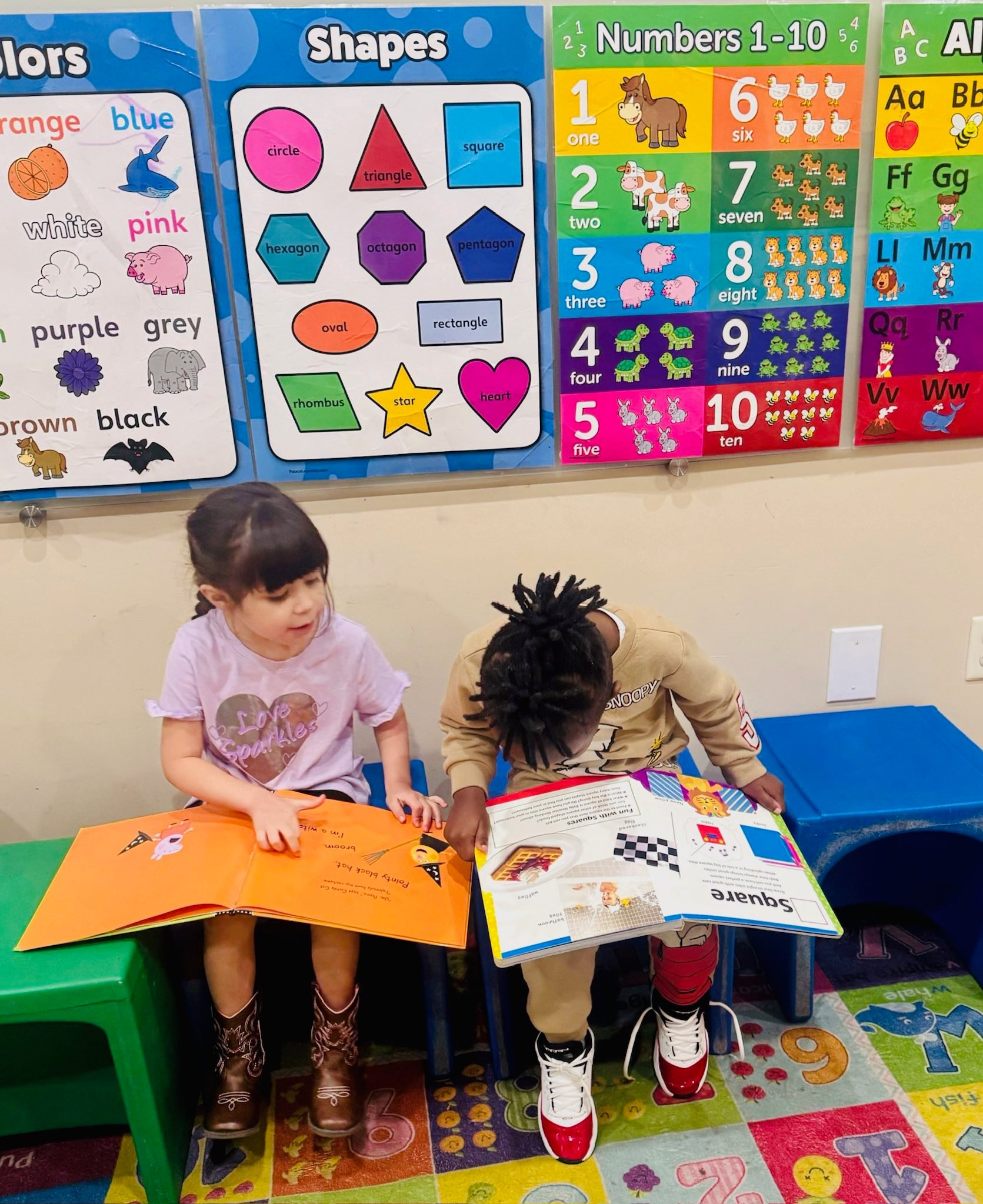 Two children reading books at a colorful classroom table with educational posters on multiplication, shapes, and numbers on the wall behind them.