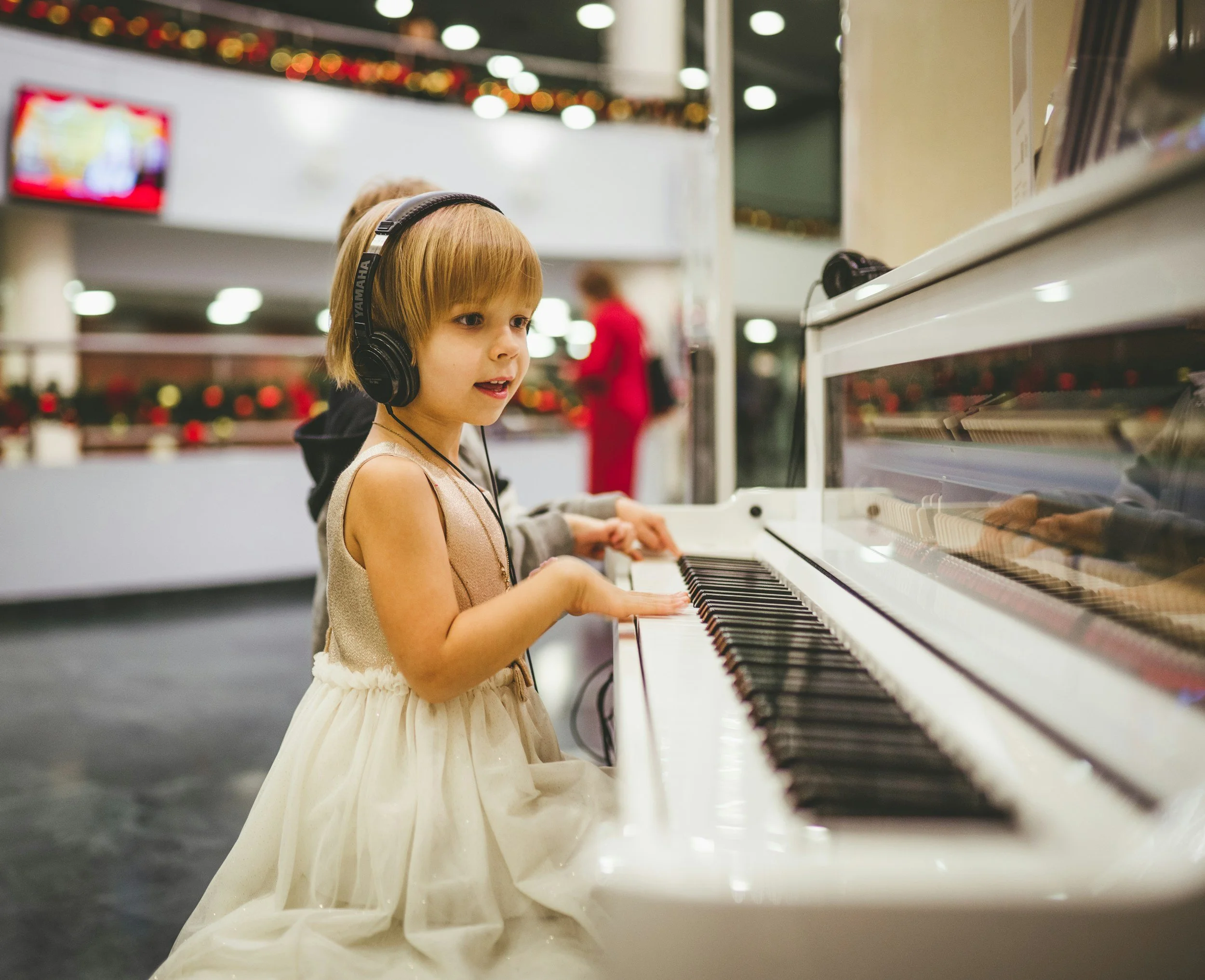 A young girl with short blonde hair wearing a beige dress and black headphones playing an electric piano in a shopping mall.