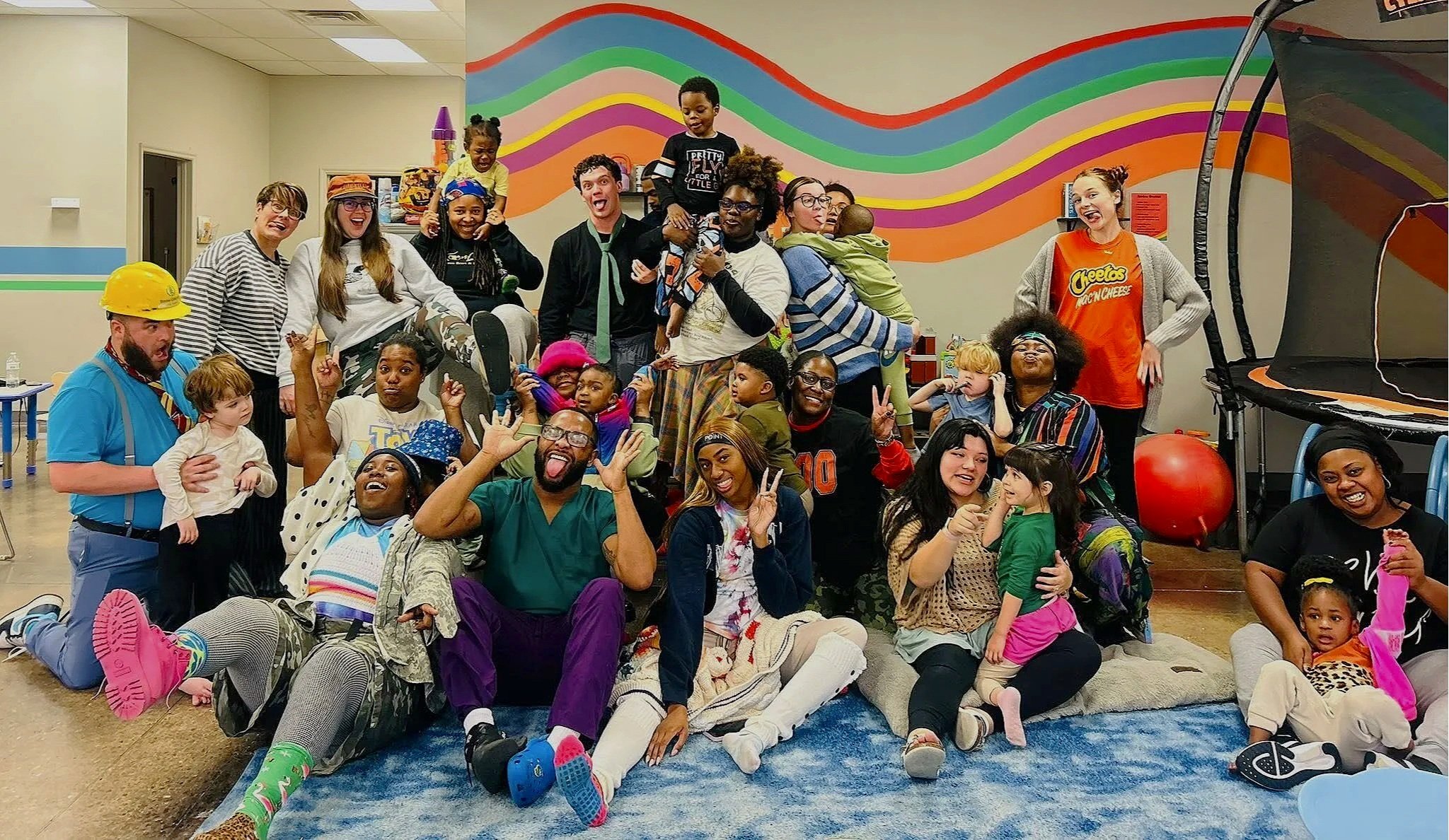 Group of children and adults posing together in a colorful indoor setting, some making funny faces, with a mural of rainbow-colored waves on the wall in the background.