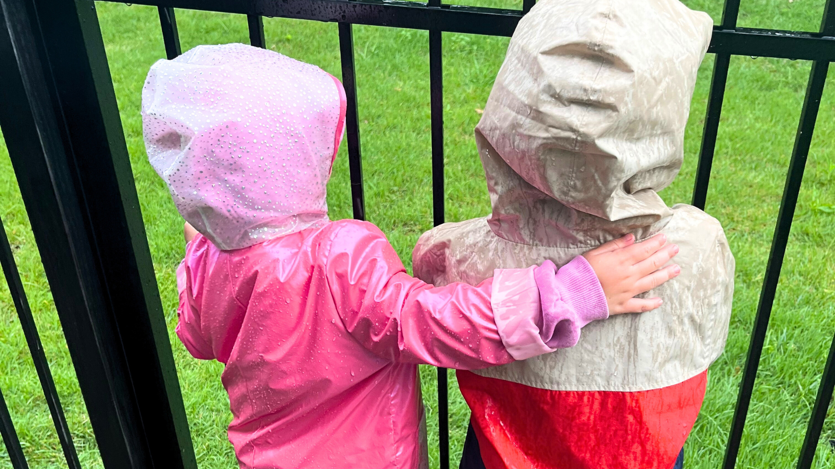 Two children wearing raincoats and hoods, standing behind a black metal fence and touching each other, on a grassy area during rainy weather.