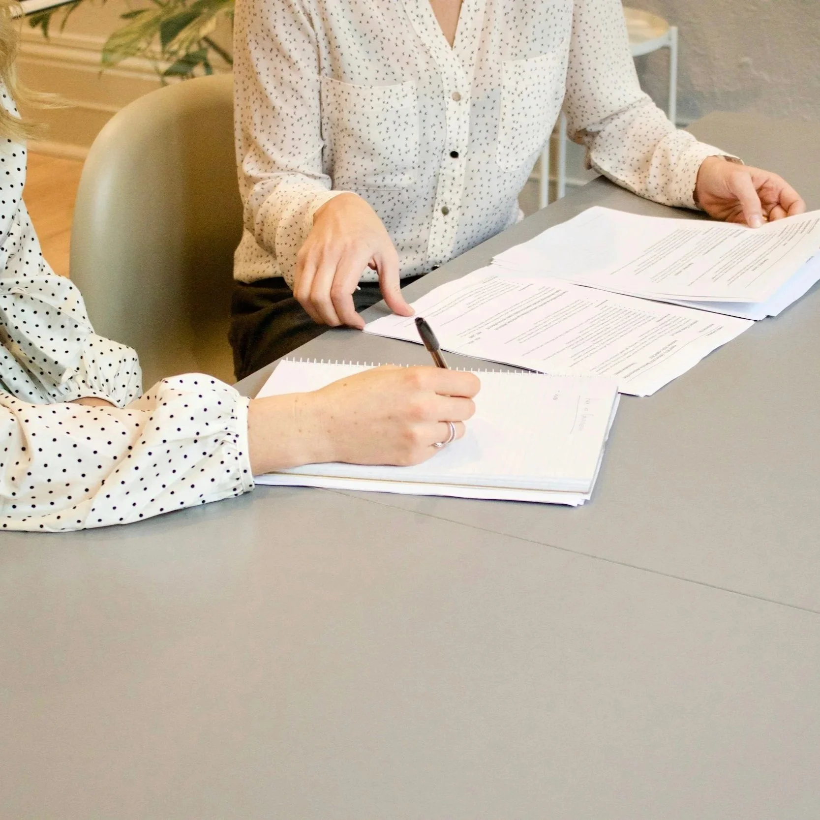 Two women sitting at a desk with papers, one is taking notes and the other is pointing at documents.