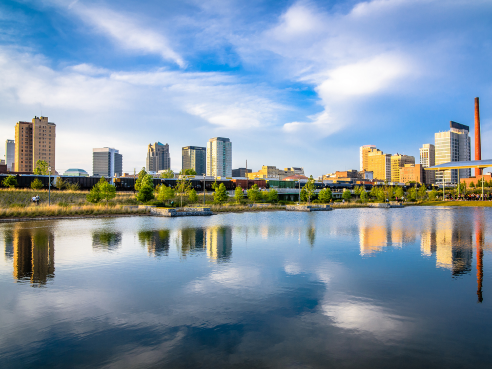 City skyline with modern buildings reflected in a river on a bright, partly cloudy day.