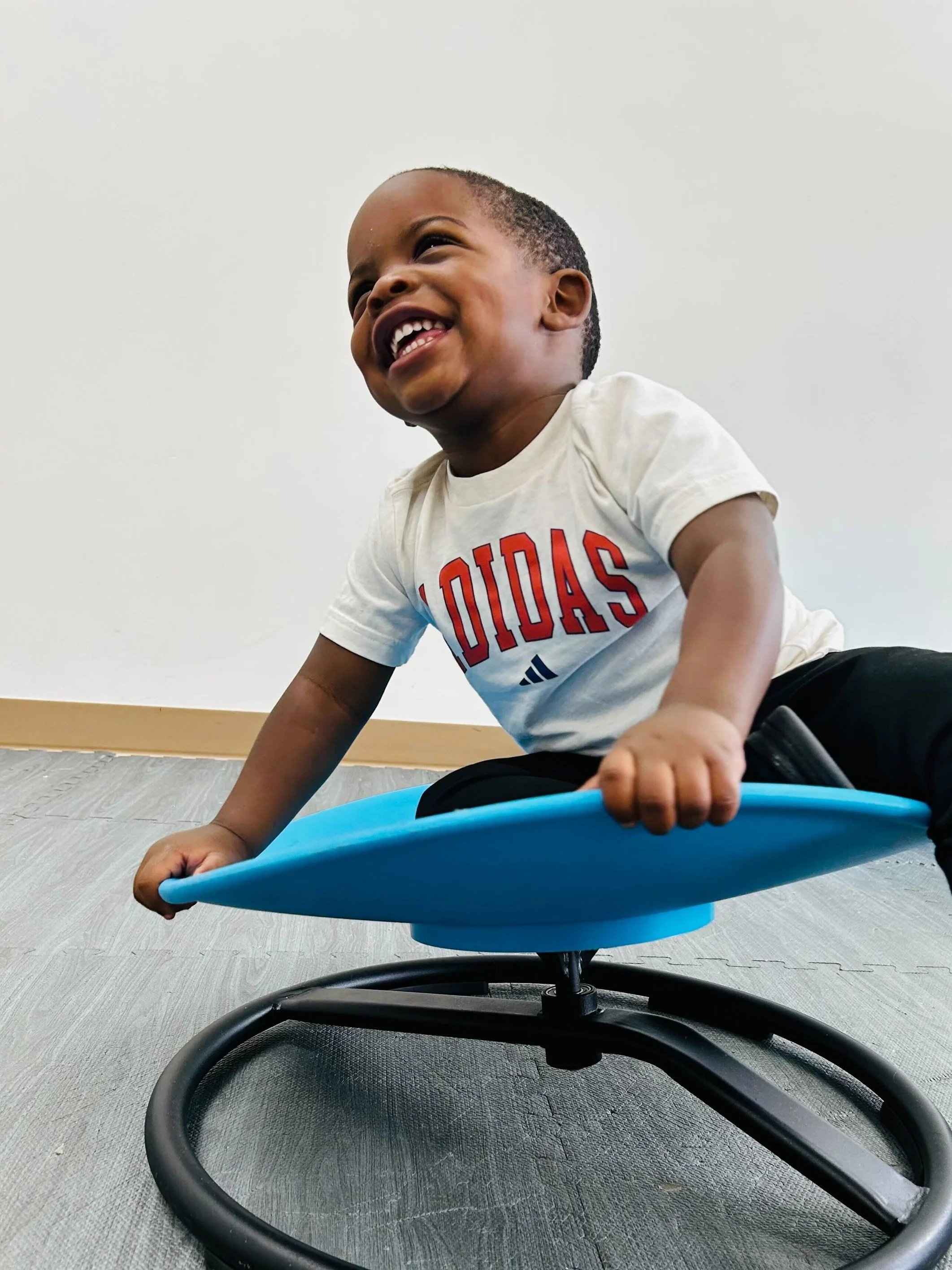 A young boy laughing joyfully while crawling on the floor, holding a blue circular balance trainer with a soccer ball patterned border.