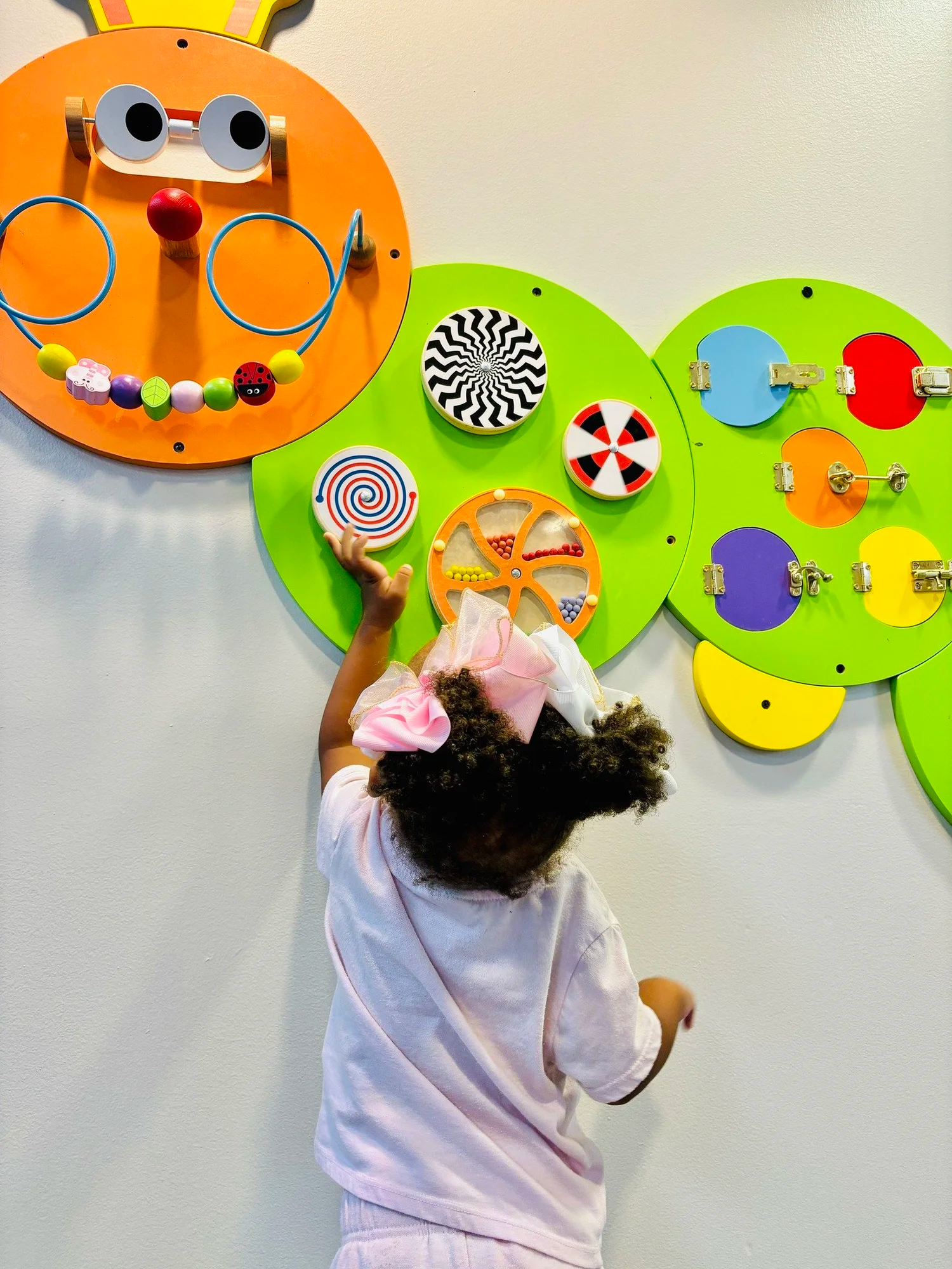 A young girl with curly hair wearing a pink bow and white pajamas stands in front of a colorful wall decoration and points at a spiral pattern on a circular board.