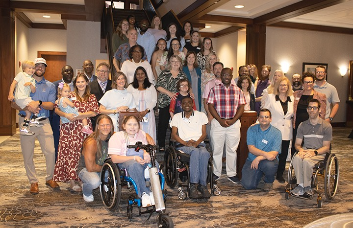 Group photo of diverse individuals, including people in wheelchairs, children, and adults, gathered indoors in a hotel or conference center setting.