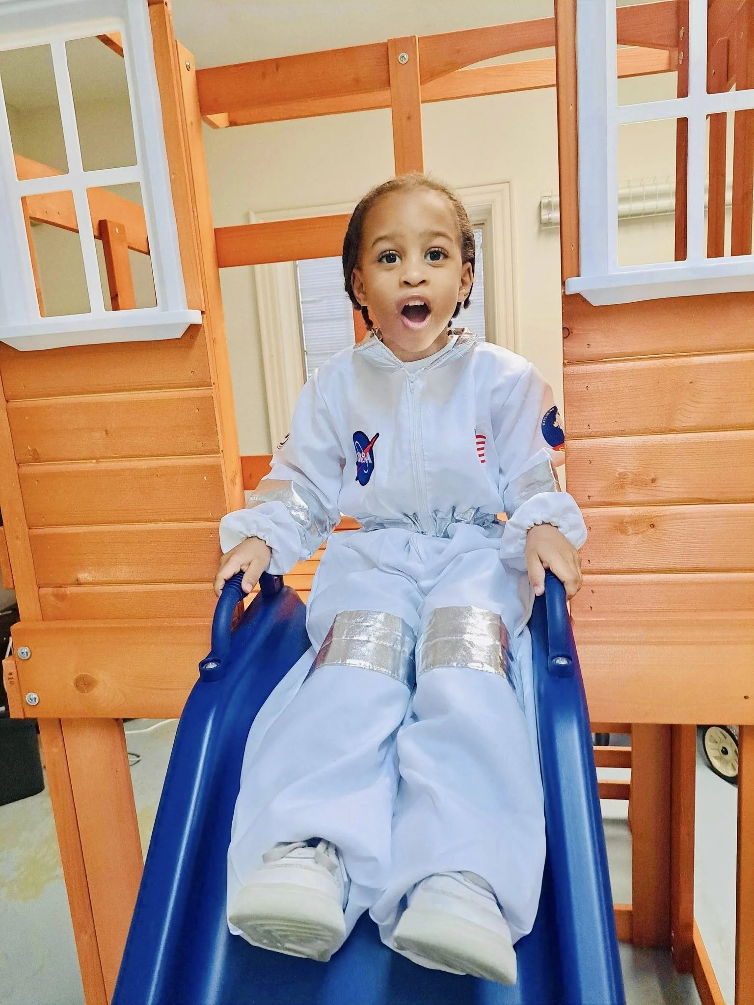 A young child on a blue slide, wearing a white NASA spacesuit costume, with an orange wooden play structure in the background.