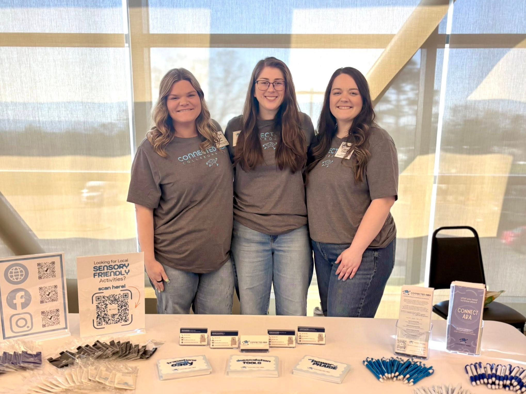 Three women standing behind a table with informational materials, promotional items, and QR codes, at an indoor event with large windows and a wooden ceiling.
