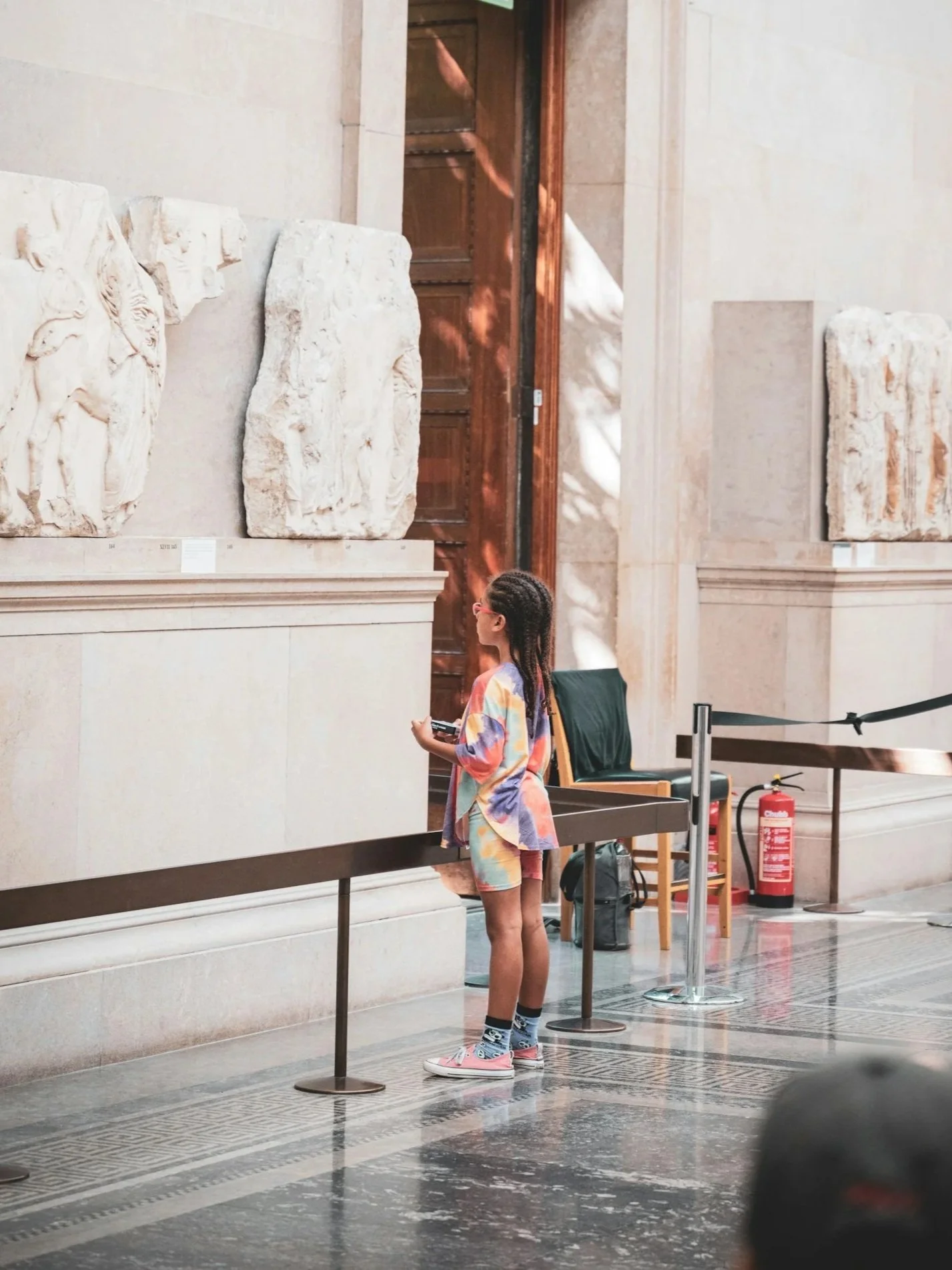 A young girl with braided hair, wearing colorful clothes, sneakers, and socks, standing in a museum, looking at ancient marble relief sculptures displayed on a wall.