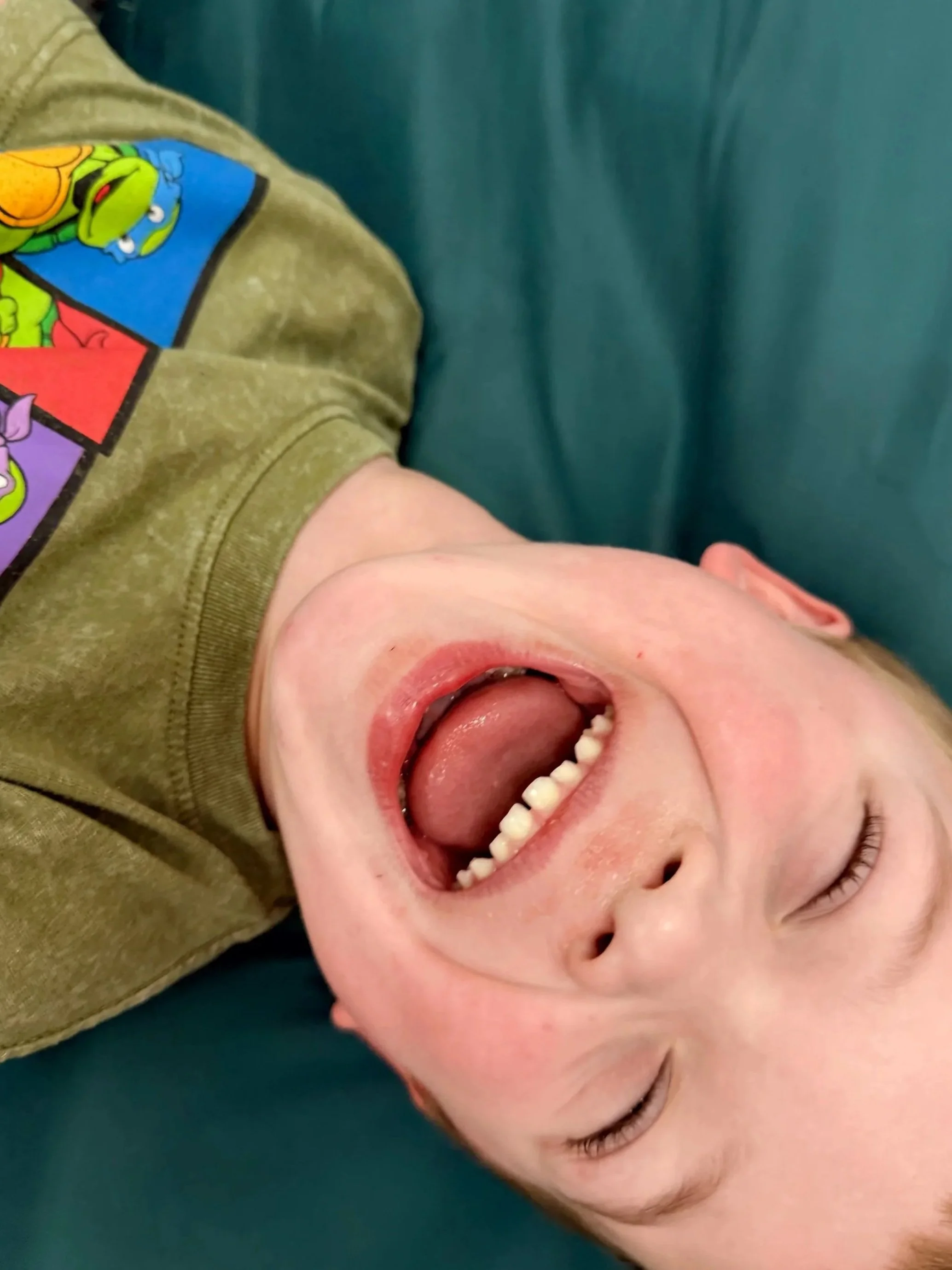 A child lying down, smiling with eyes closed, mouth wide open, showing teeth, wearing a green shirt with Teenage Mutant Ninja Turtles patch.