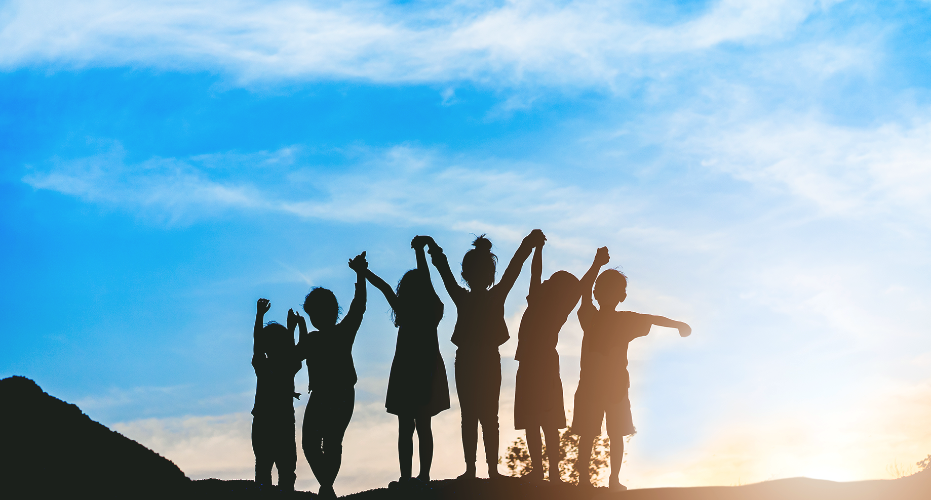 Silhouettes of children holding hands and raising arms joyfully against a sunset sky with clouds.