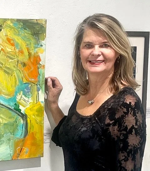 A woman in a black lace dress standing next to an abstract painting with yellow, green, orange, and blue strokes at an art gallery.