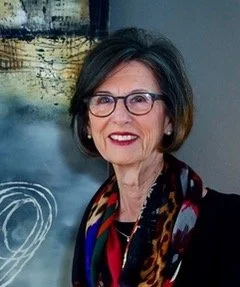 Portrait of an older woman with short dark hair, wearing glasses, a dark blazer, and a colorful scarf, smiling in front of a water feature.