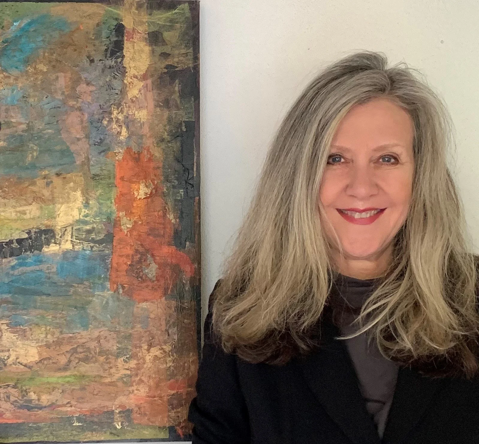 A smiling woman with long, wavy gray hair stands next to an abstract painting on a white wall.