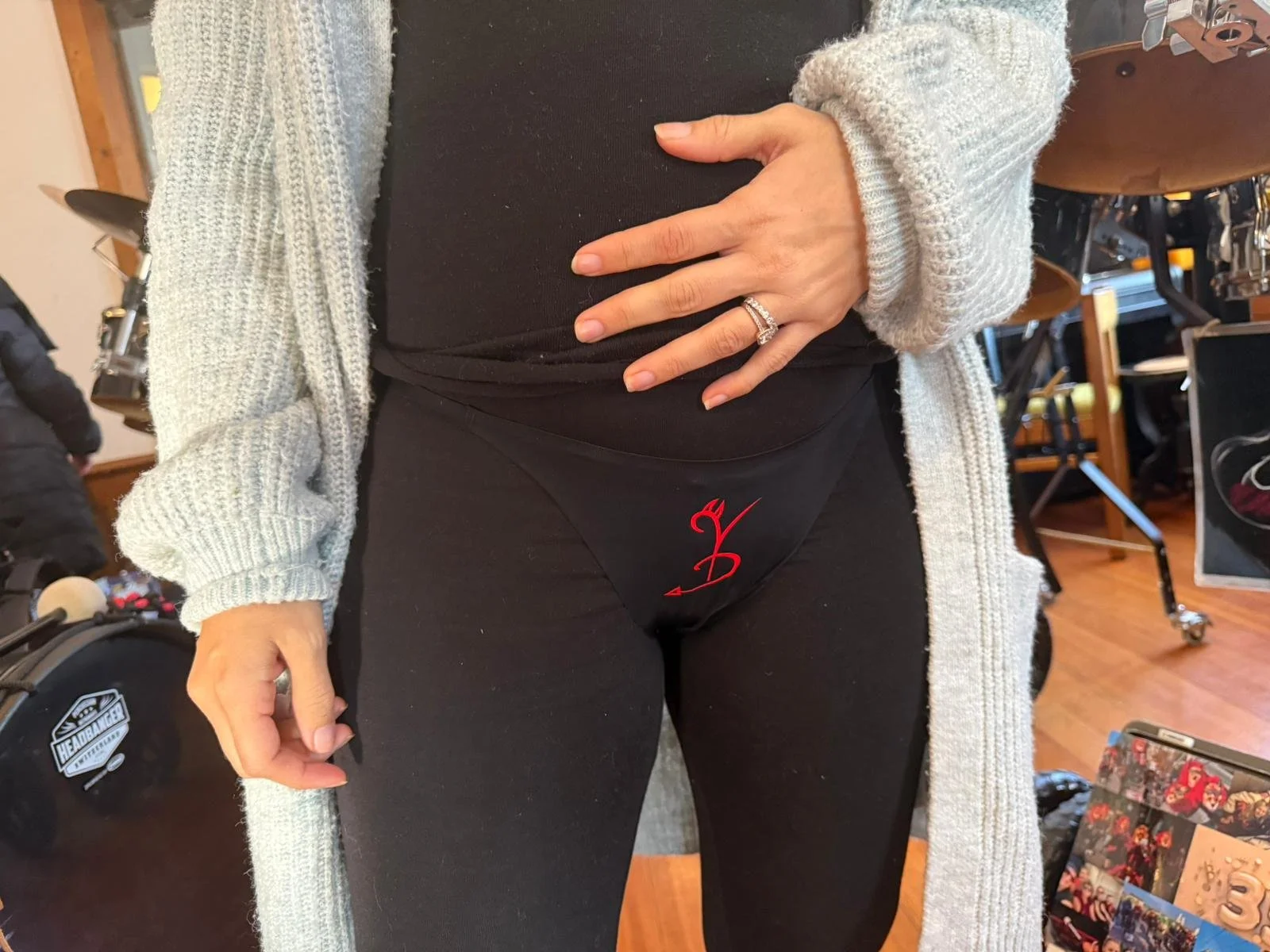 Nahaufnahme eines weiblichen Bauches mit schwarzem T-Shirt und grauem Strickmantel, das eine Hand mit Ehering auf den Bauch legt, im Hintergrund Musik- und Geräteausstattung in einem Raum.