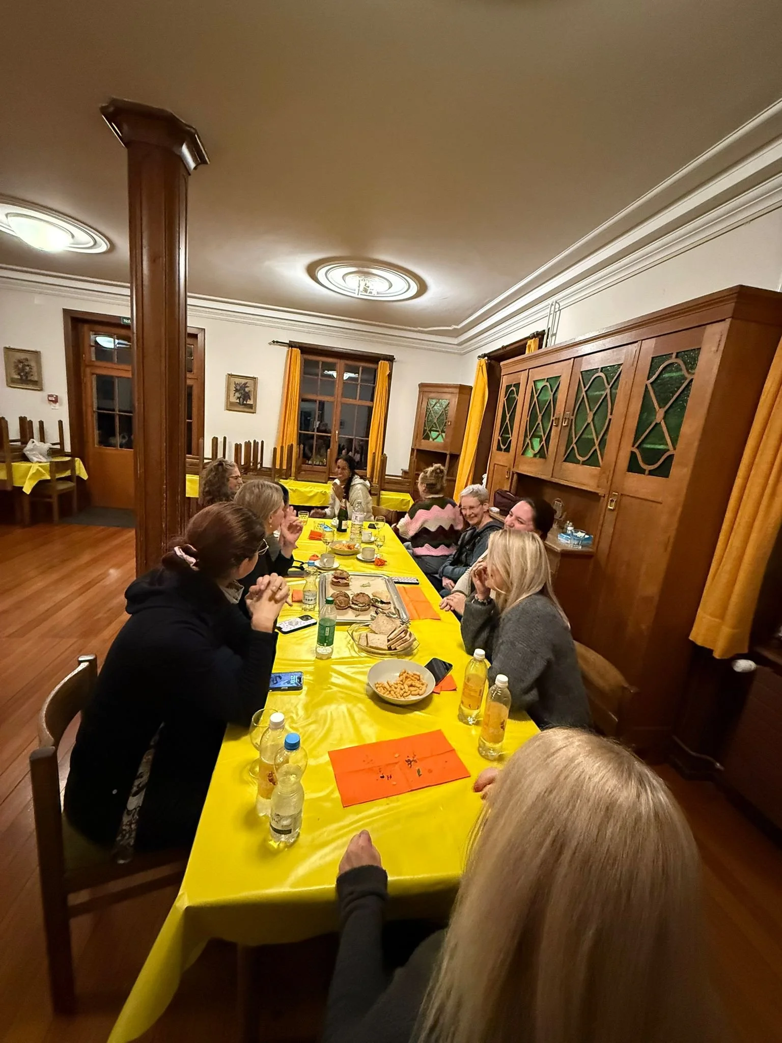 Mehrere Menschen sitzen an einem länglichen Tisch in einem festlich dekorierten Raum, essen und trinken. Der Tisch ist mit einer gelben Tischdecke bedeckt, es gibt Getränke und Snacks. Im Hintergrund sind Fenster mit gelben Vorhängen und Holzschränke