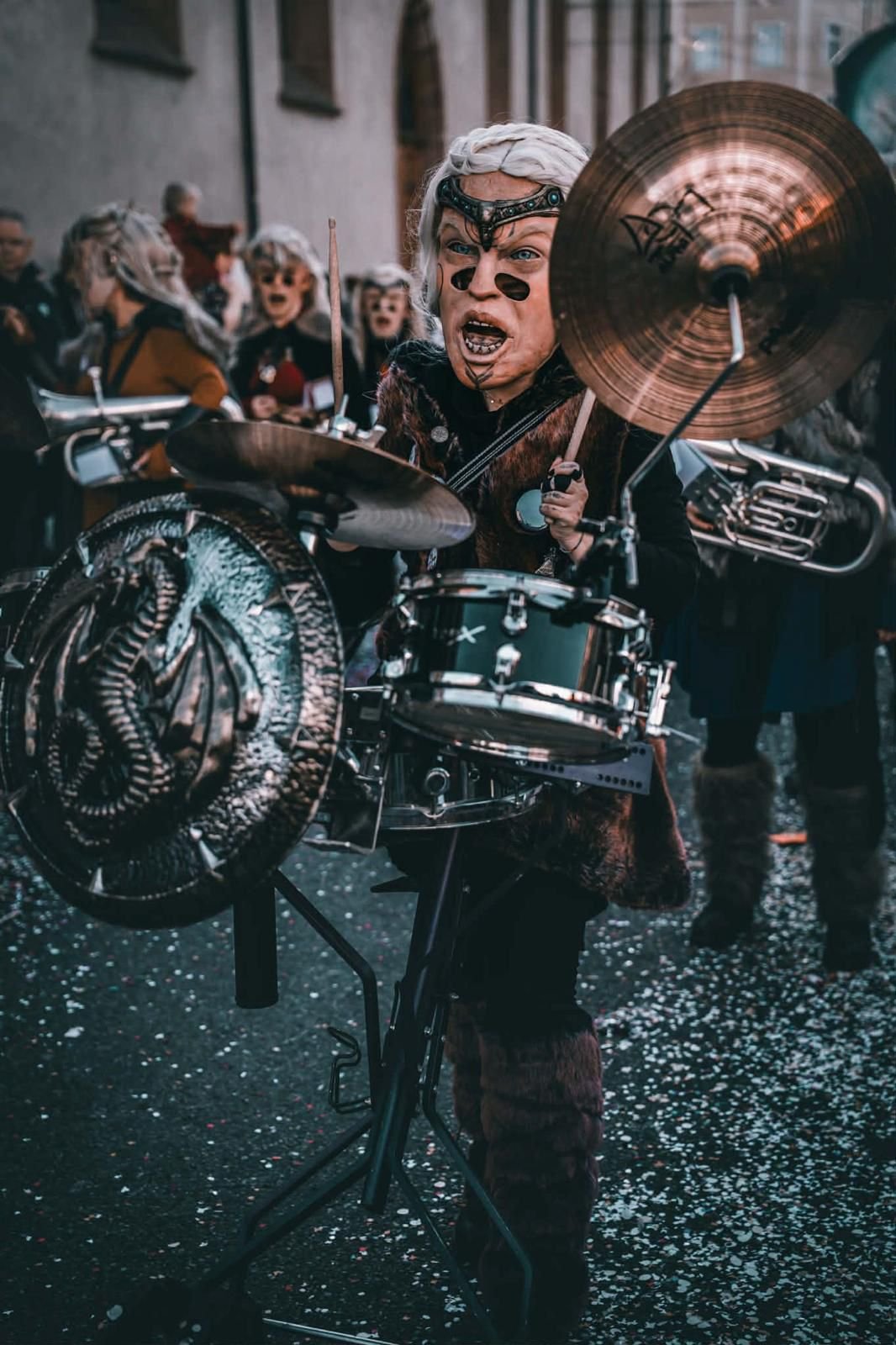 Eine Person in einem Kostüm mit einem maskenähnlichen Gesicht spielt Schlagzeug bei einer Parade oder Feier, umgeben von anderen in ähnlichen Kostümen, auf einer Straße mit Konfetti.
