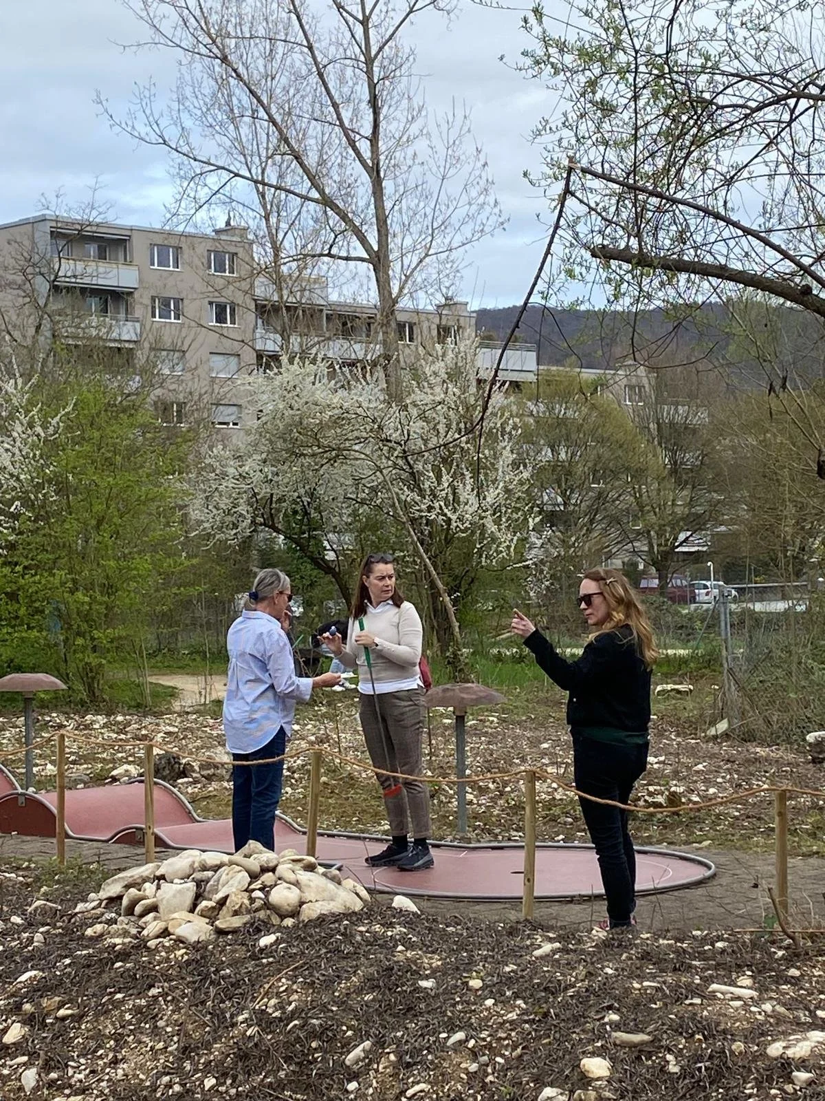 Drei Frauen stehen auf einem kleinen Spielplatz im Freien, mit vielen Bäumen und an einem Gebäude in der Hintergrund bei bewölktem Himmel.