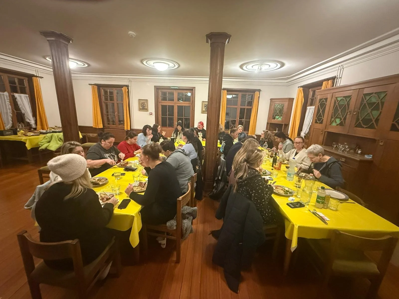 Mehrere Menschen sitzen an Tischen in einem Raum, essen und trinken. Der Raum hat Holzwände, große Fenster mit Gelbvorhängen und braune Holzfußböden.
