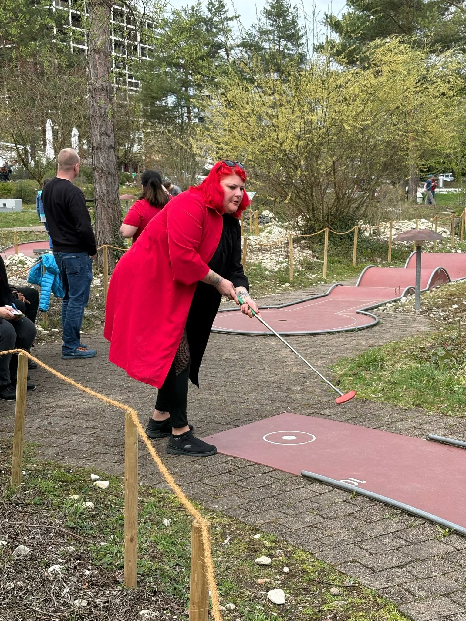 Eine Frau mit roten Haaren und rotem Mantel spielt Minigolf auf einer Outdoor-Anlage.