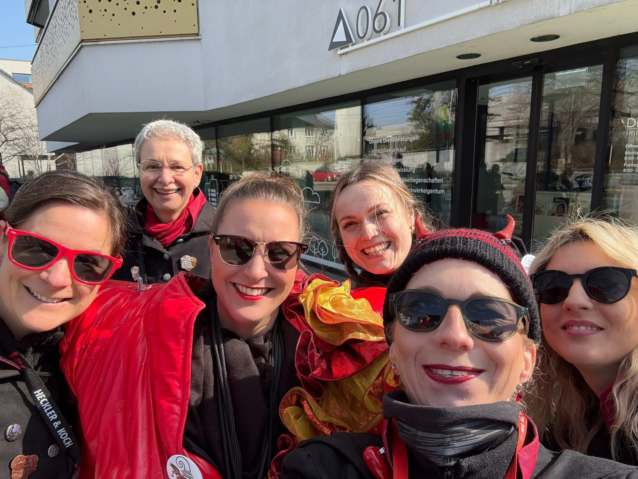 Sieben lachende Frauen mit Sonnenbrillen und bunten Outfits vor einem Geschäftsgebäude.