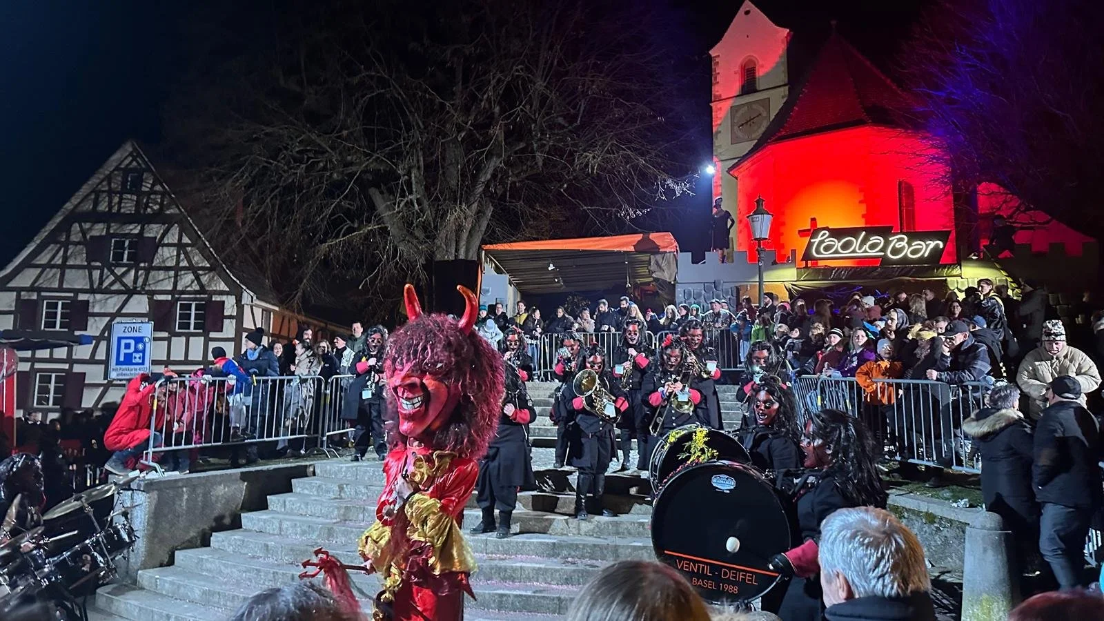Menschen in Kostümen und Masken bei einer nächtlichen Veranstaltung auf den Treppen vor einem Gebäude, mit einem Teufelskostüm in der Nähe, im Hintergrund ein Schilder mit 'Tool Bar' und eine Kirche, viele Zuschauer.