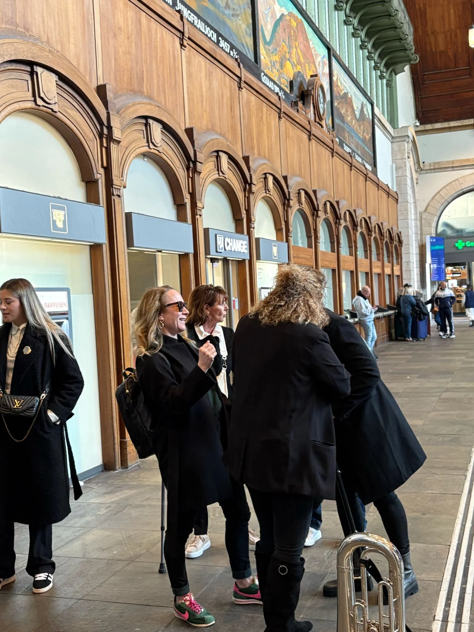 Menschen warten in einem Bahnhof, einige unterhalten sich, andere stehen in der Nähe eines Schalterbereichs.