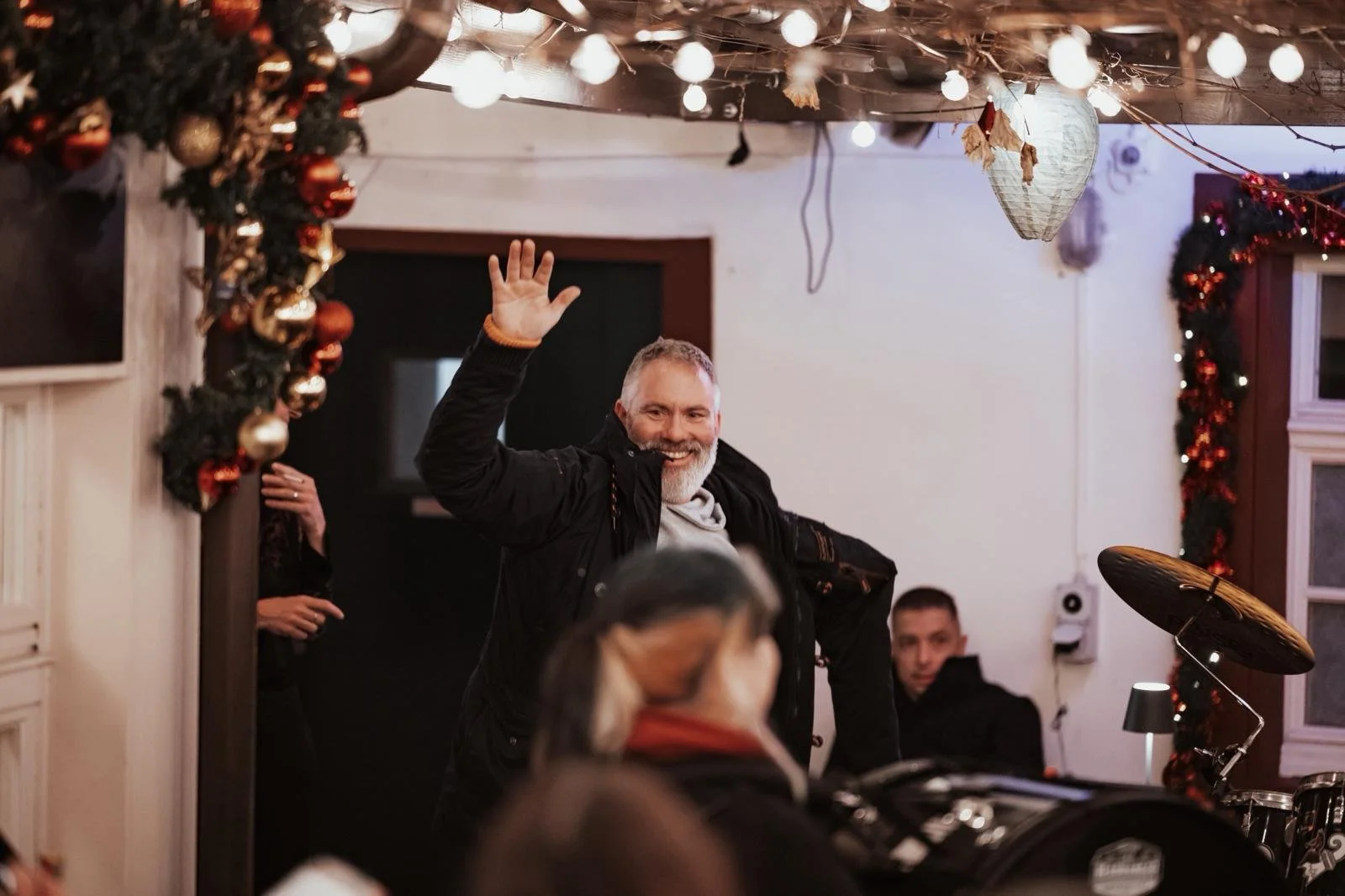 Ein Mann mit grauem Haar und Bart, der lächelnd in einem festlich geschmückten Raum winkt.