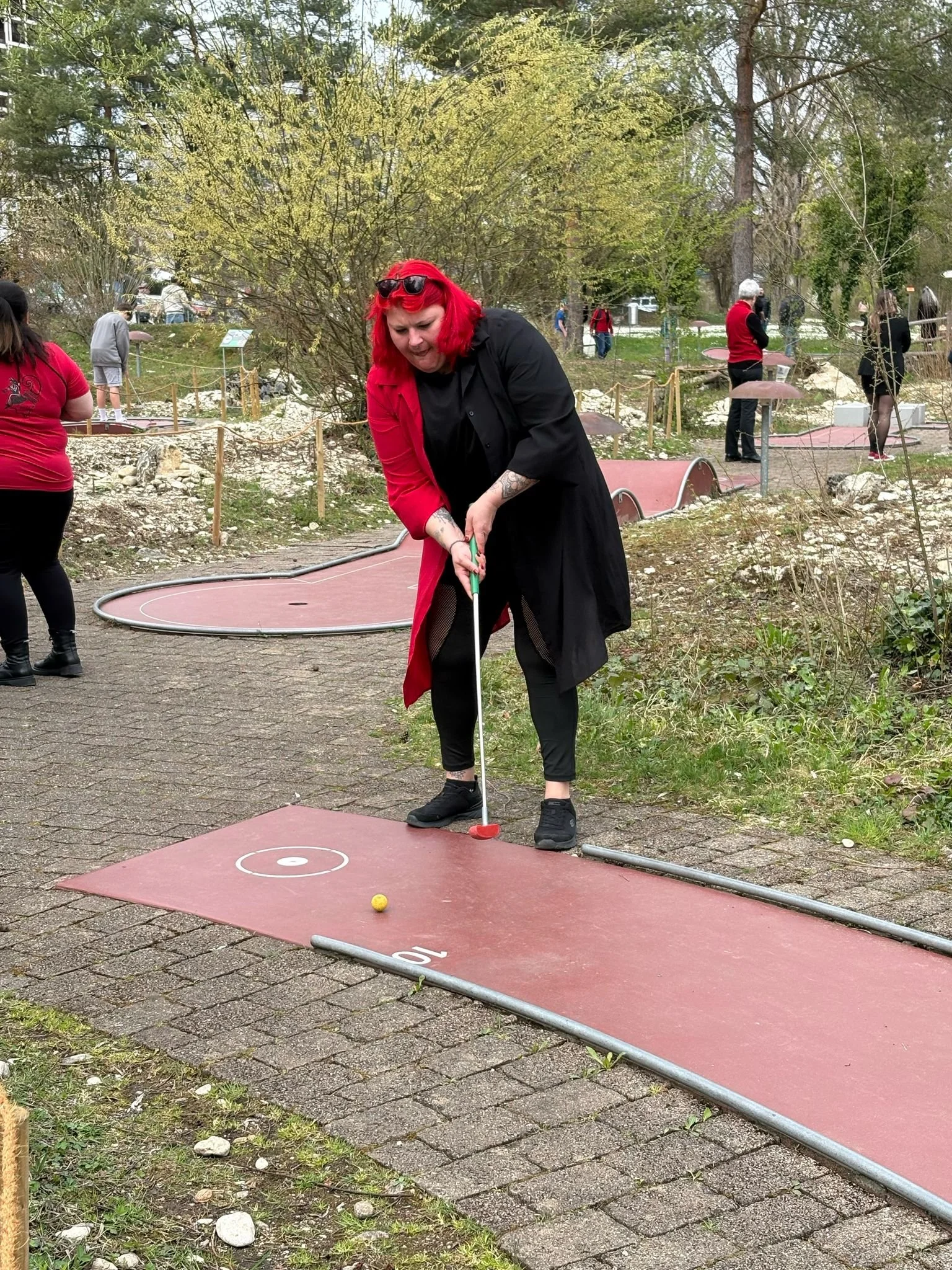 Eine Frau mit roten Haaren und dunkler Kleidung spielt Minigolf auf einer Outdoor-Bahn. Es sind weitere Personen im Hintergrund, die ebenfalls Minigolf spielen.