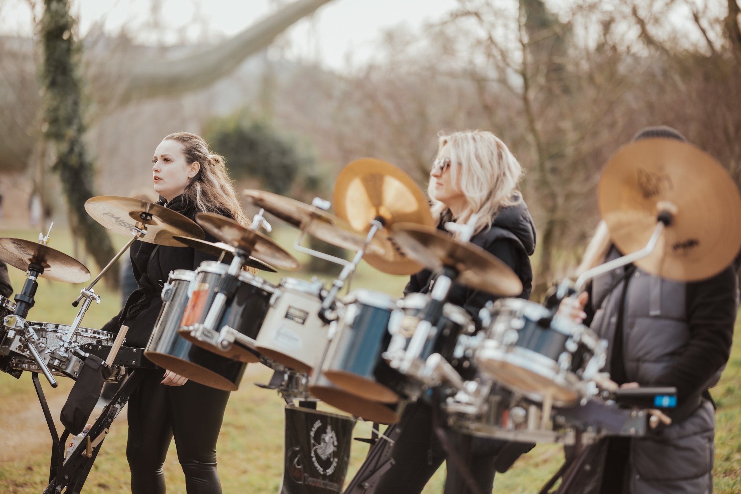 Ein Mädchen mit langen, blonden Haaren spielt Schlagzeug in einem Park, umgeben von Bäumen, mit anderen Musikern.