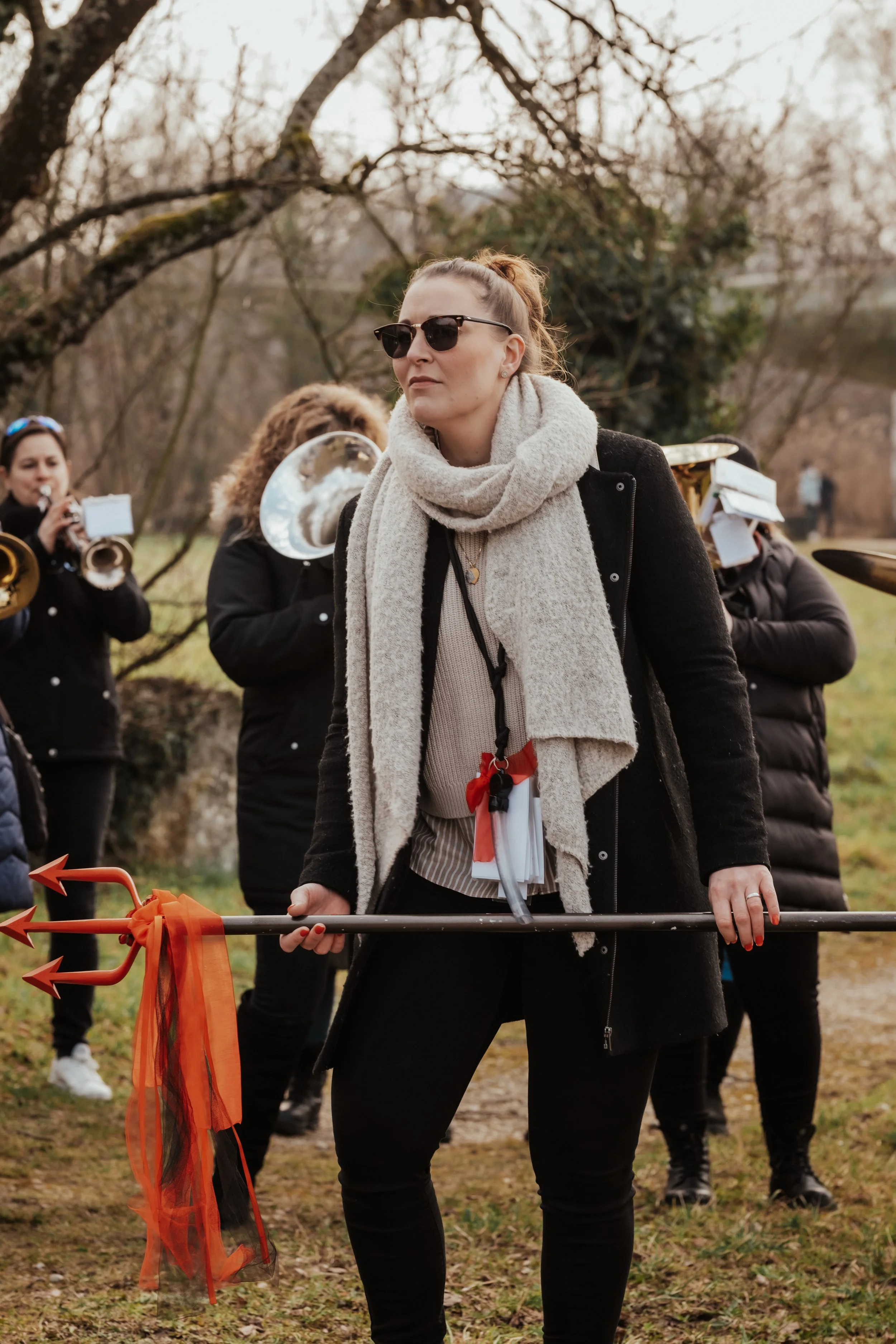Eine Frau mit Sonnenbrille, grauem Schal und schwarzen Kleidern hält einen roten Keulen-Stab mit Tüll vor einem Hintergrund von Musikern, die Blasinstrumente spielen, in einem outdoor Park bei kühlem Wetter.