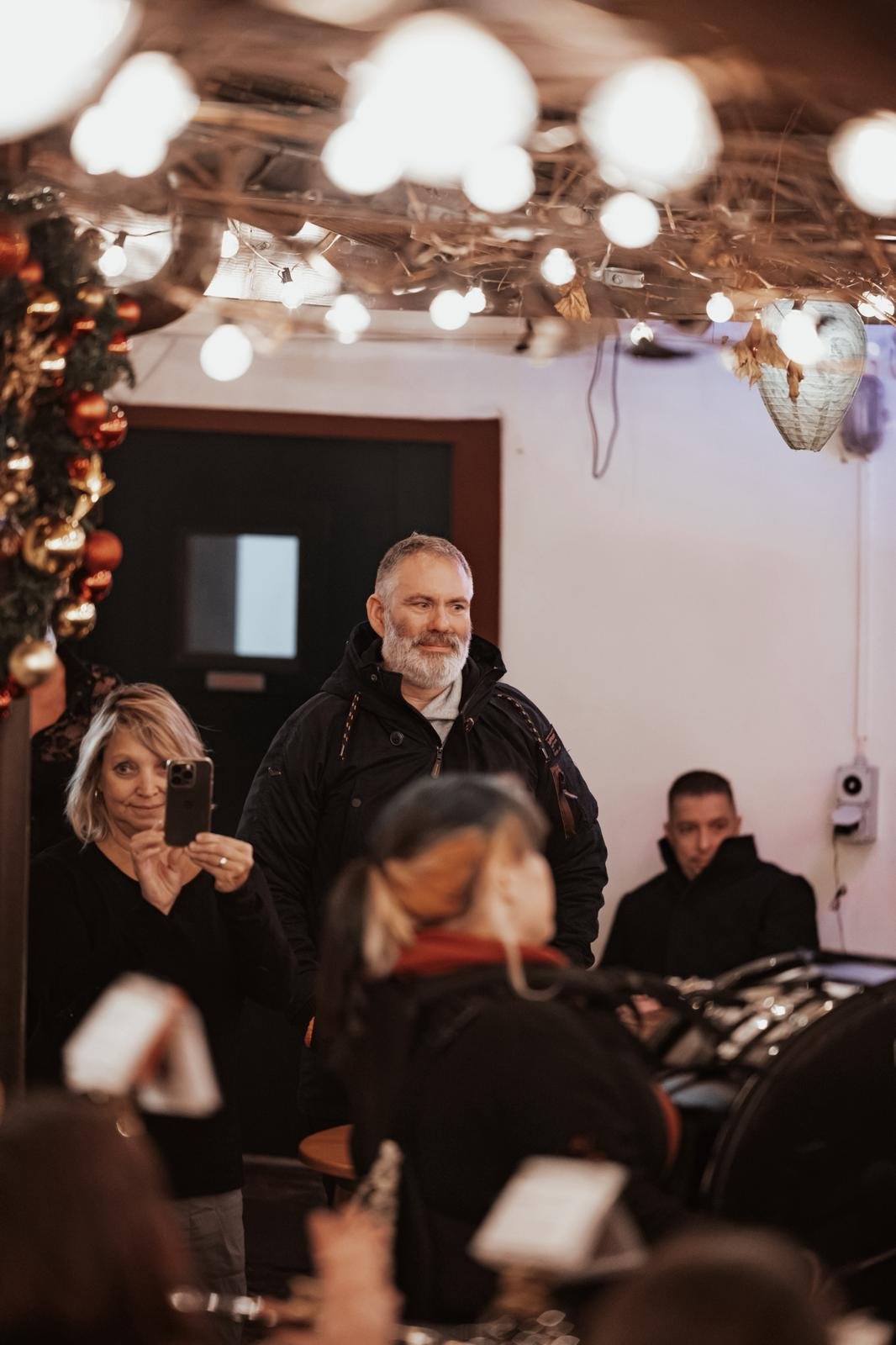 Gruppe von Menschen in einem festlich dekorierten Raum, einige machen Fotos, während andere zuschauen, Weihnachtsdekorationen und Lichter sind sichtbar.