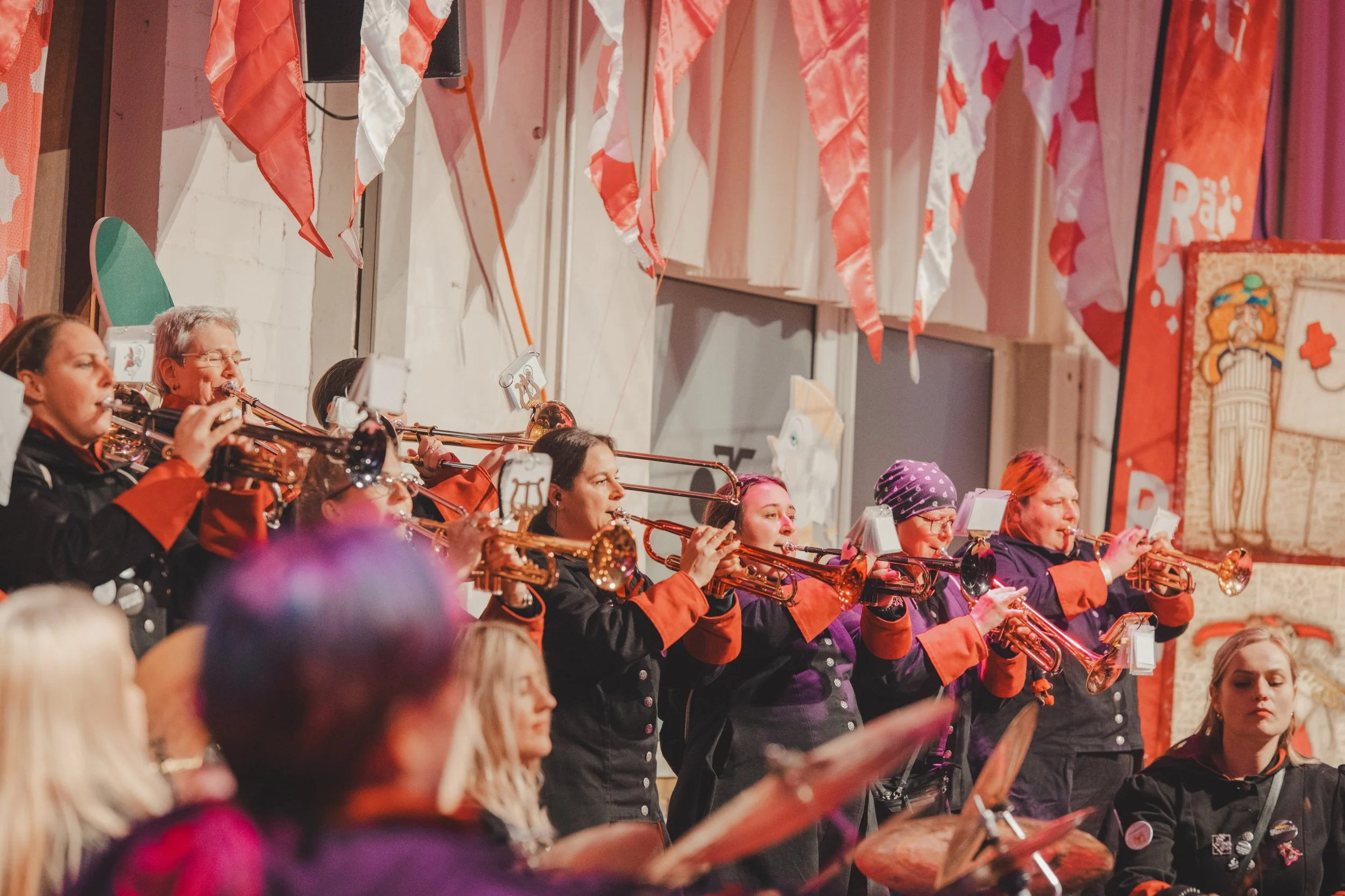 Mehrere Musiker spielen Flöte auf einer Bühne bei einer Veranstaltung. Sie tragen schwarz-orange Kleidung, einige tragen Brillen und einen trägt ein violettes Stirnband. Im Hintergrund sind rote und weiße Fahnen und Dekorationen sichtbar.