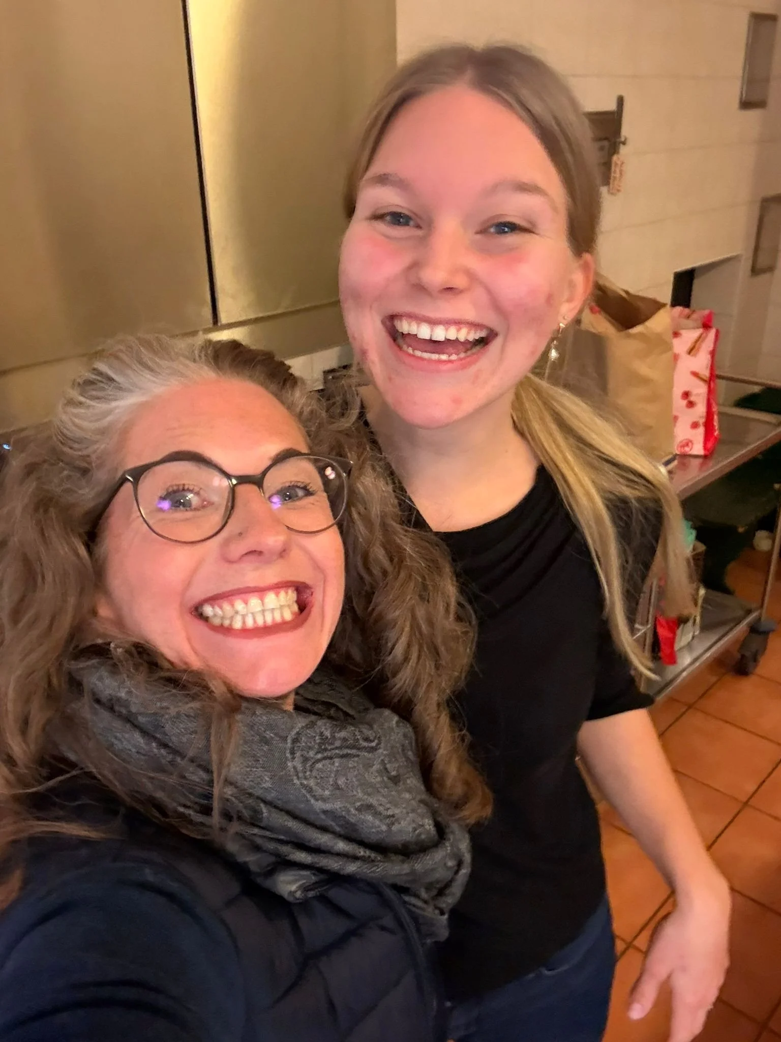 Zwei lachende Frauen, eine mit Brille und grauem Schal, die andere ohne Brille, in einem Raum mit goldener Wand. Sie posieren für ein Selfie.