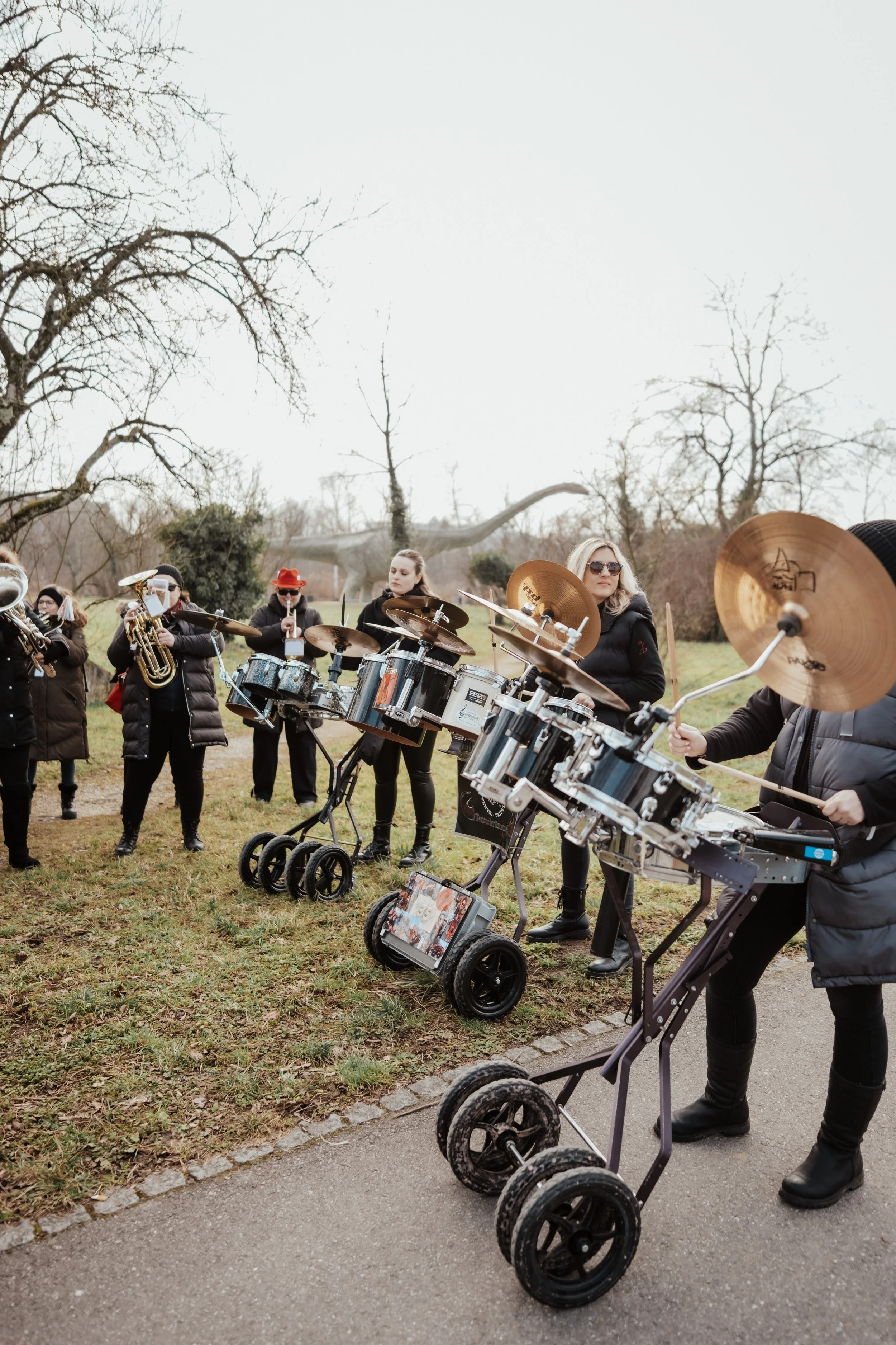 Gruppe von Musikern, die im Freien auf einem Rasen spielen, unter Bäumen mit mehrstöckigen Dinosaurier-Dekoration im Hintergrund.