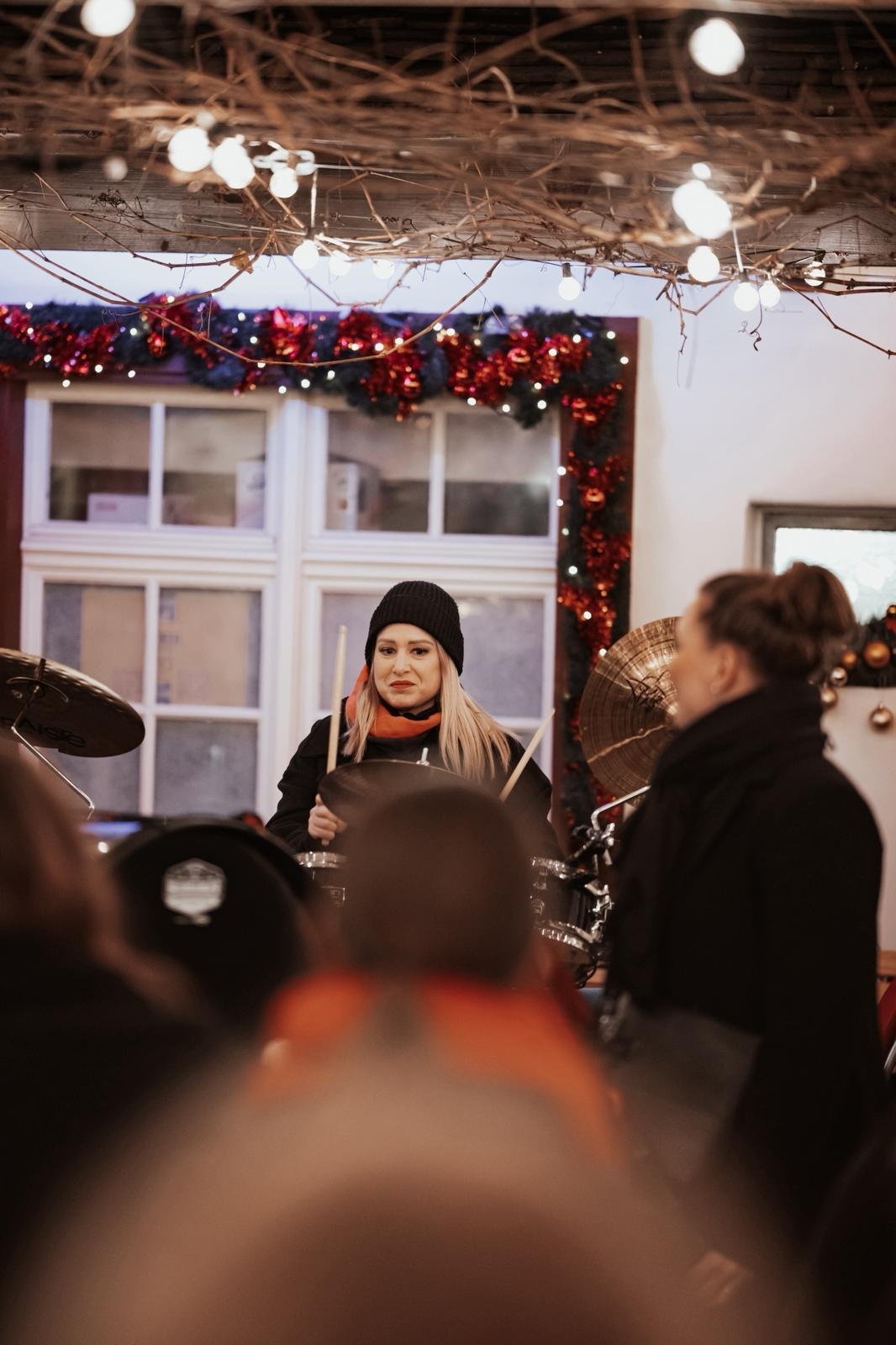 Eine junge Frau mit blondem Haar trägt eine schwarze Mütze und hält Trommelstöcke, während sie in einer festlich geschmückten Umgebung spielt. Im Vordergrund sind die Köpfe anderer Personen, die offenbar ein Konzert oder eine Feier besuchen.