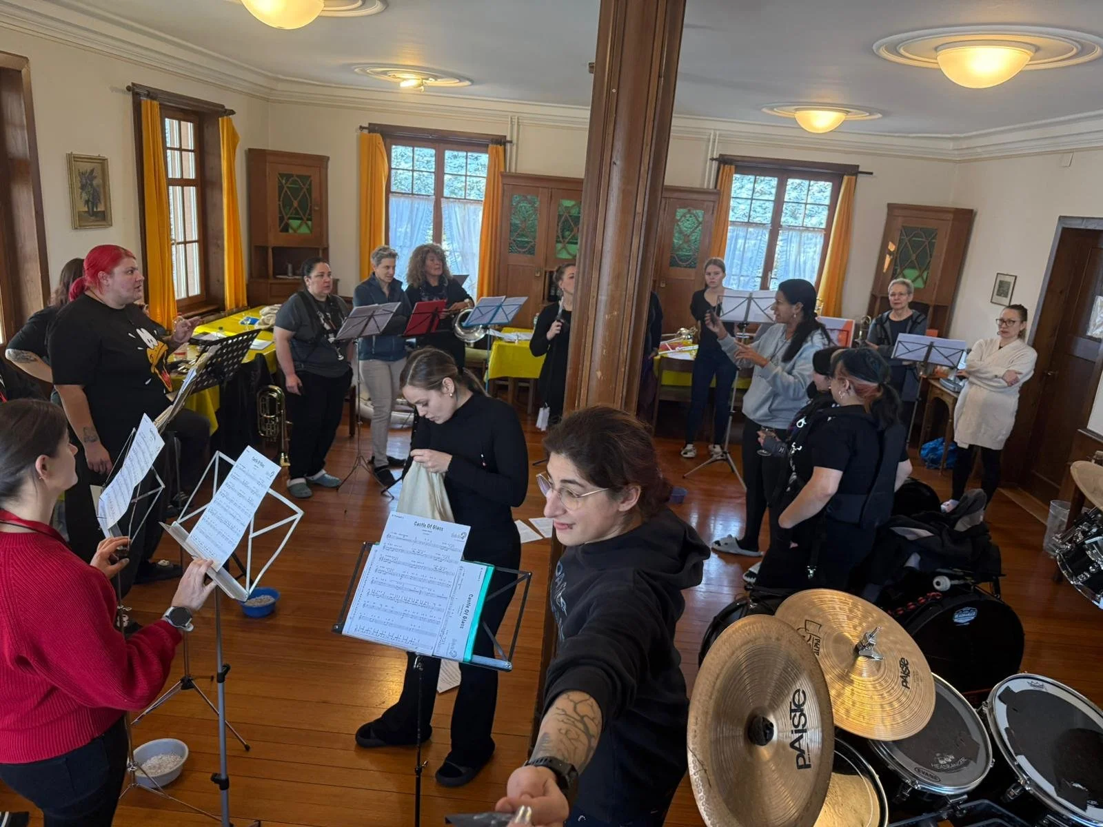 Eine Frau posiert vor einer Holzsäule in einem Raum mit mehreren Musikerinnen und Musikern, die sich auf eine Probe oder ein Konzert vorbereiten.