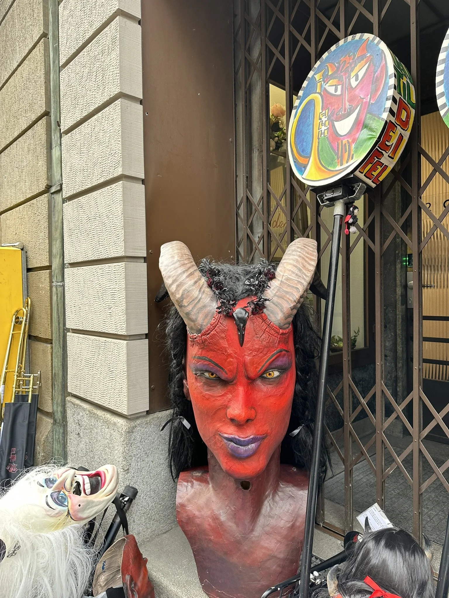 Eine große Teufelsmaske mit roten Gesicht, schwarzen Augen und Hörnern steht vor einem Gebäude, umgeben von weiteren Masken und Figuren.
