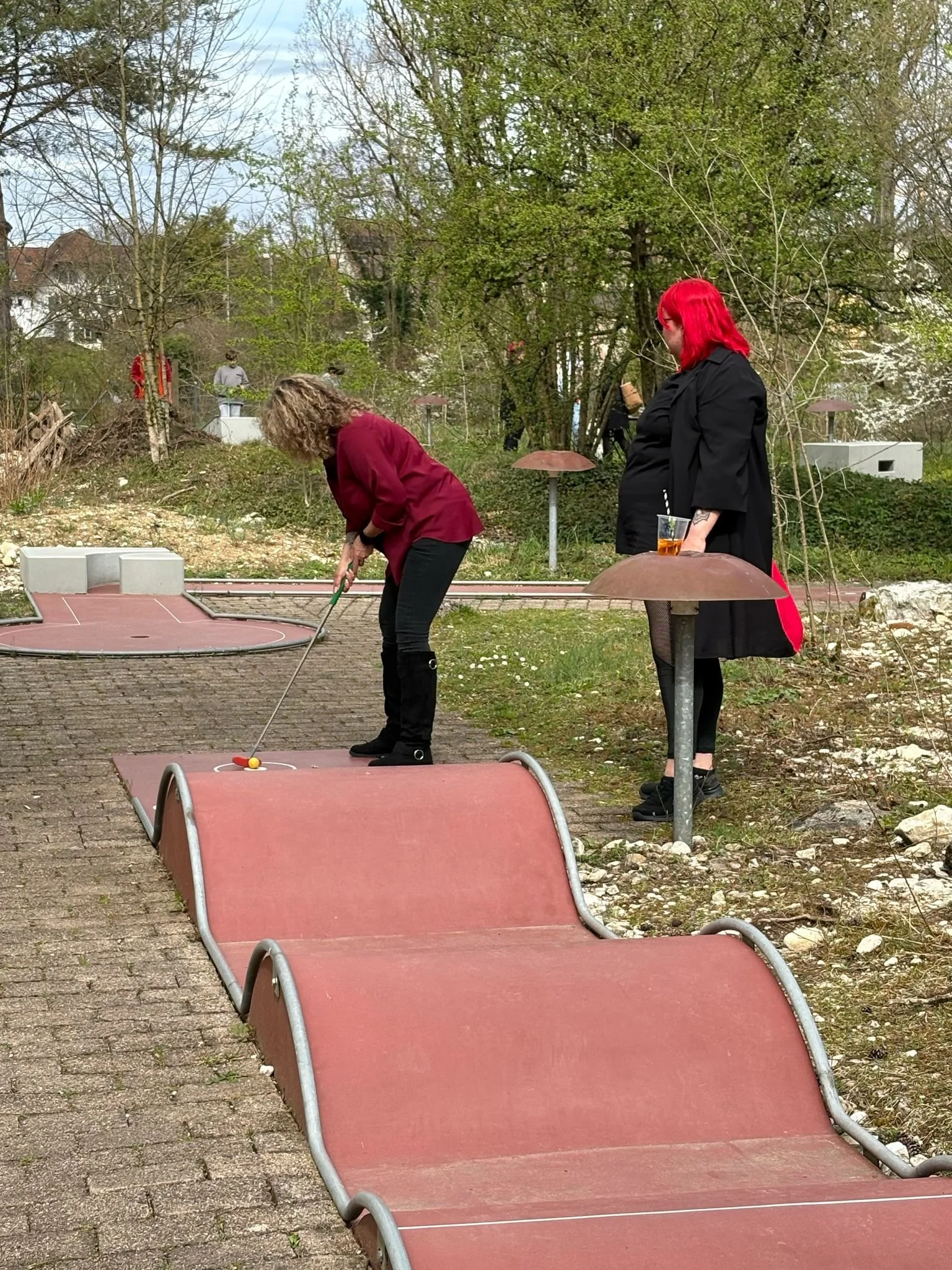 Zwei Frauen stehen auf einem Mini-Golfplatz. Die eine Frau bereitet einen Schlag vor, während die andere neben einem Getränkehalter steht und einen Drink hält. Im Hintergrund sind Bäume und einige Mini-Golf-Stationen sichtbar.
