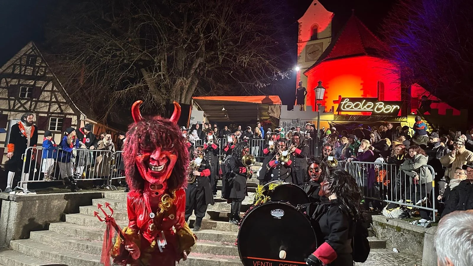 Traditionelles Fasnachtskostüm mit Teufelsmaske und Hörner vor einer Fasnachtsumzugsgruppe, die auf dem Treppenaufgang einer historischen Kirche in einer Altstadt- Umgebung auftritt, bei nächtlichem Fest in Deutschland.