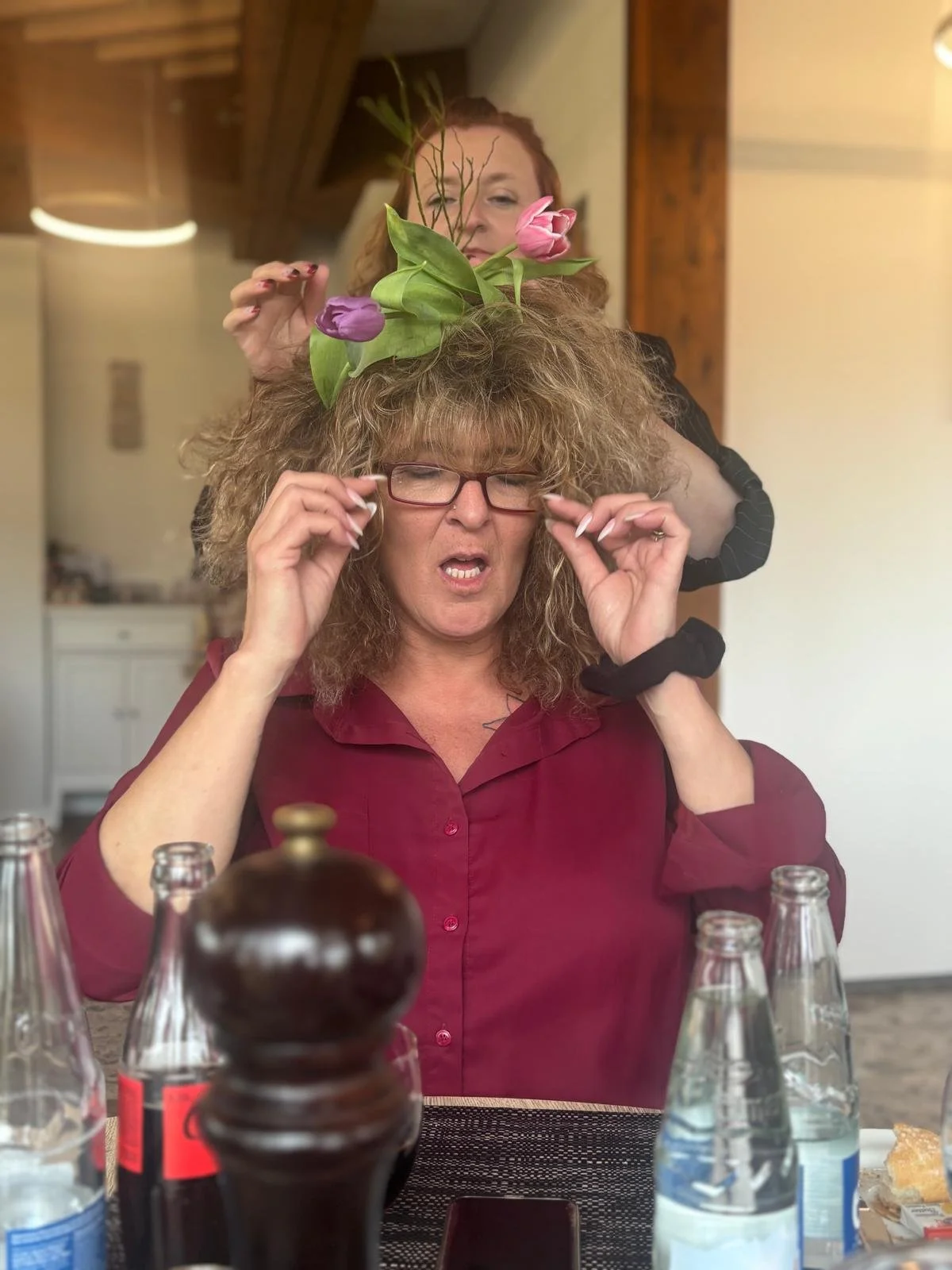 Frau mit großen lockigen Haaren trägt eine Brille, während eine andere Person Blumen im Haar der Frau steckt, im Hintergrund holzverkleidete Wand.