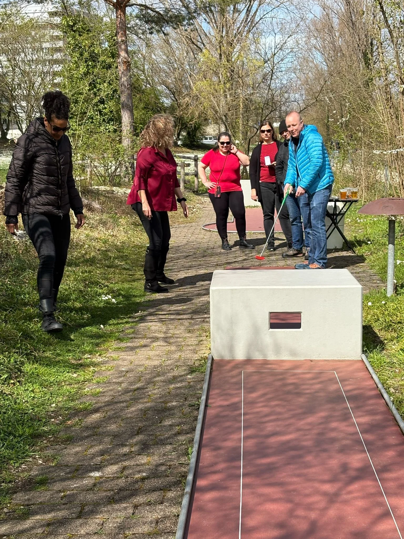 Eine Gruppe von sechs Personen spielt Minigolf im Freien an einem sonnigen Tag. Eine Person schlägt den Ball, während die anderen zuschauen.