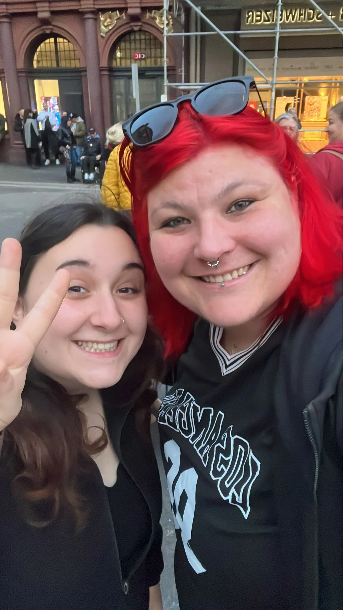 Zwei lachende Frauen machen ein Selfie auf der Straße, eine mit roten Haaren und Sonnenbrille auf dem Kopf, die andere zeigt ein Peace-Zeichen.
