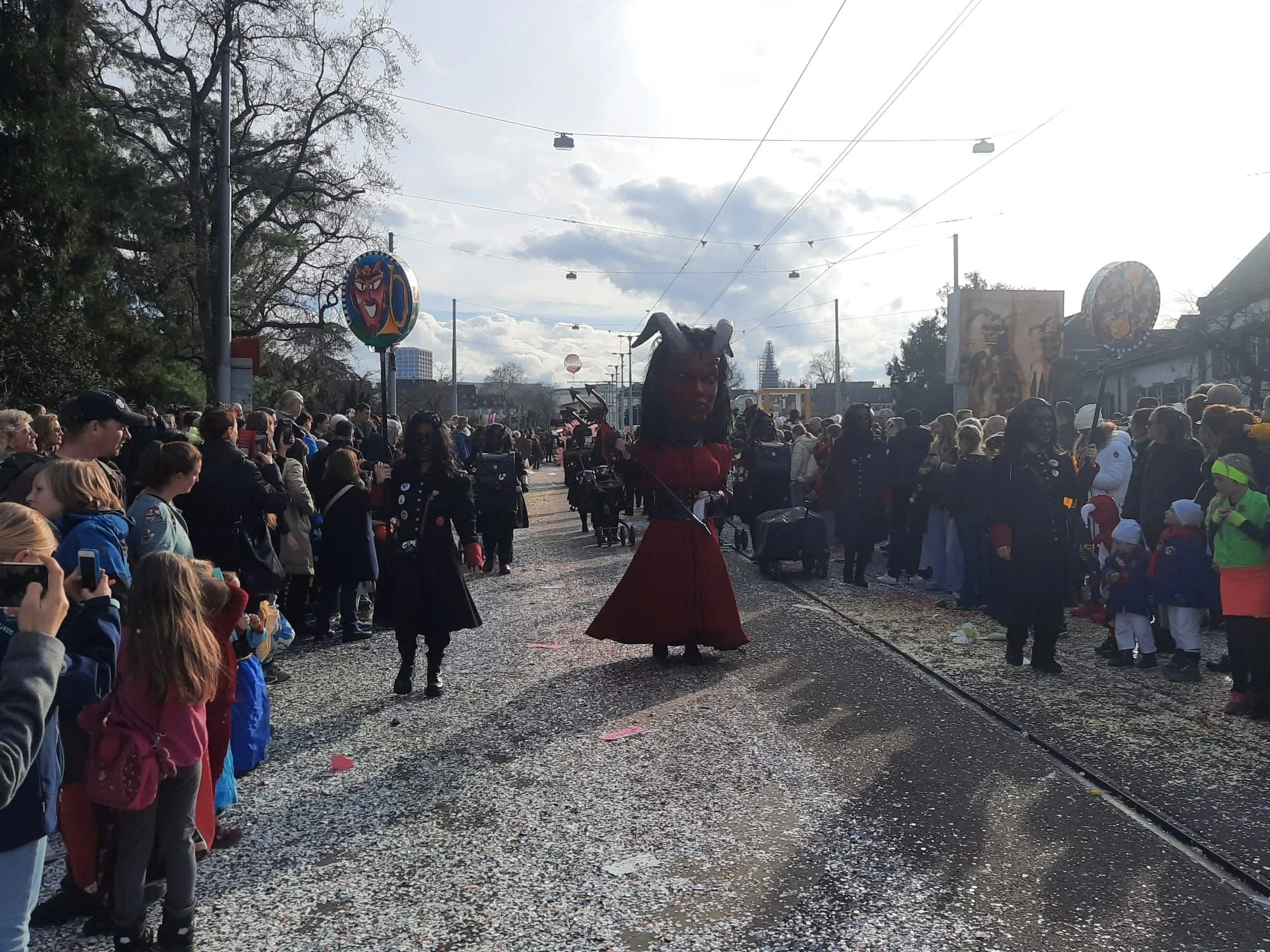 Festzug mit Menschen in Kostümen während eines Parades in einer Stadt, im Hintergrund sind Bäume, Gebäude und dekorative Figuren sichtbar.