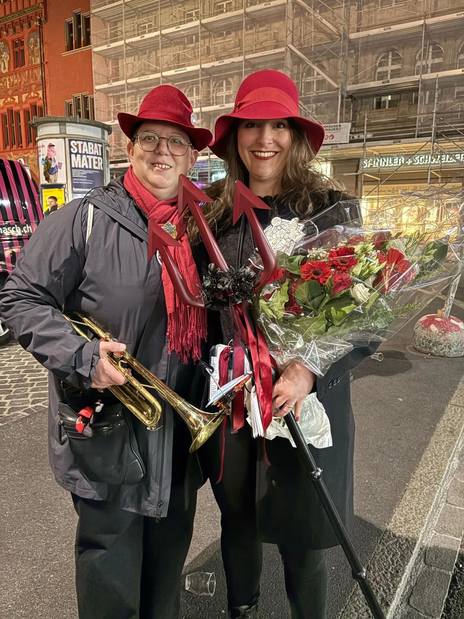 Zwei Frauen mit roten Hüten, eine trägt ein Saxophon, die andere hält einen Blumenstrauß und ein rotes Trident, beide lächeln und posieren auf der Straße bei Nacht.