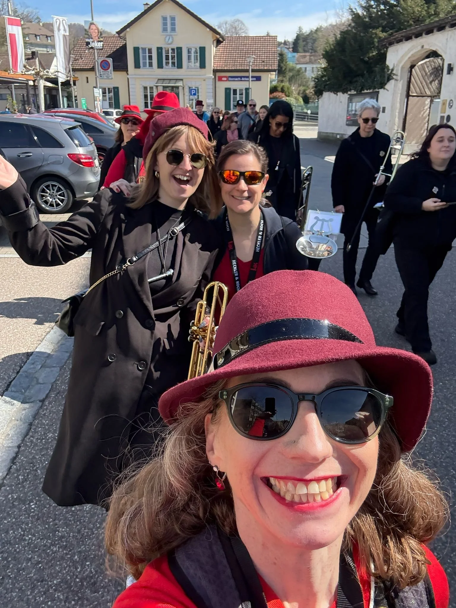 Mehrere Frauen, einige mit Musikinstrumenten, posieren im Freien in einer Stadtstraße. Sie tragen bunte Hüte und Sonnenbrillen.