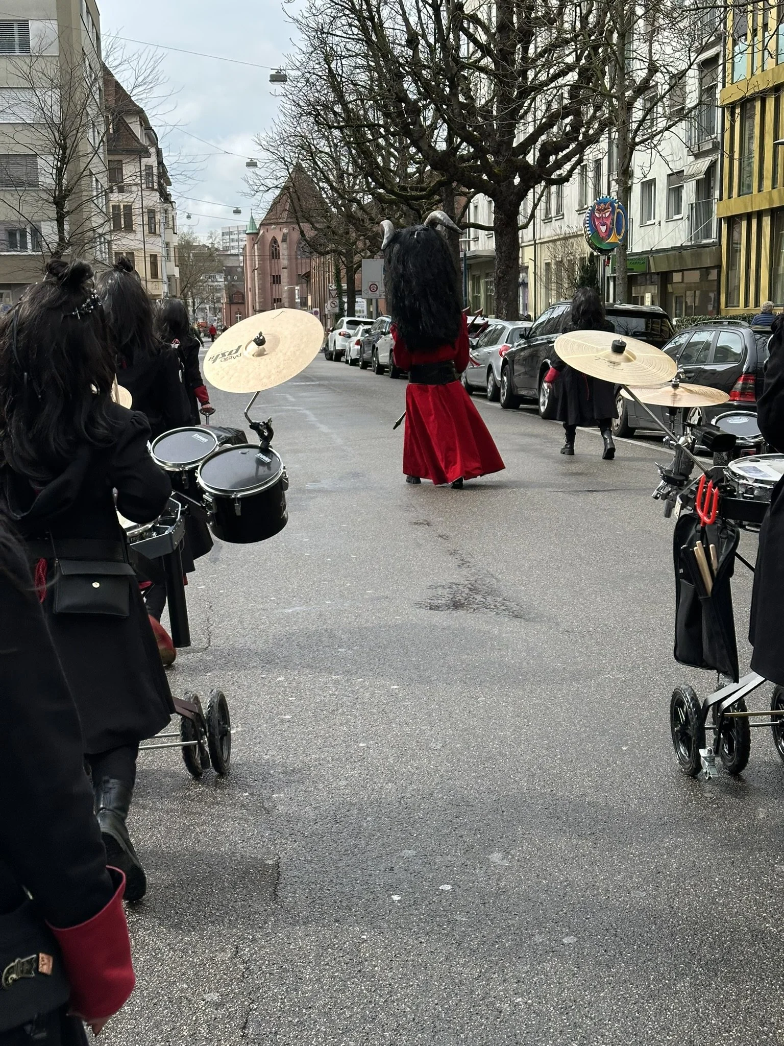 Gruppierung von Menschen in einem urbanen Street-Setting, mit einer Figur im Kostüm mit langen schwarzen Haaren und Hörnern, die auf der Straße steht, während andere Personen mit Musikinstrumenten im Vordergrund unterwegs sind.